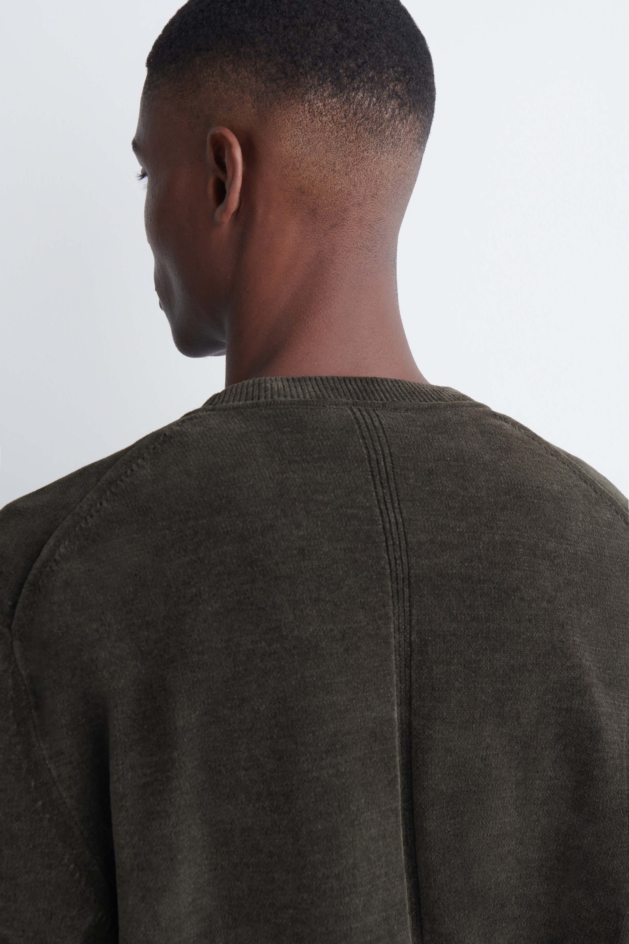 A dark olive green cotton chenille crew-neck sweater is being worn by the model, shown from the back to highlight the back design. The sweater features a textured knit, a ribbed neckline, and a vertical seam running down the center back. A dark olive green cotton chenille crew-neck sweater is being worn by the model, shown from the back to highlight the back design. The sweater features a textured knit, a ribbed neckline, and a vertical seam running down the center back.