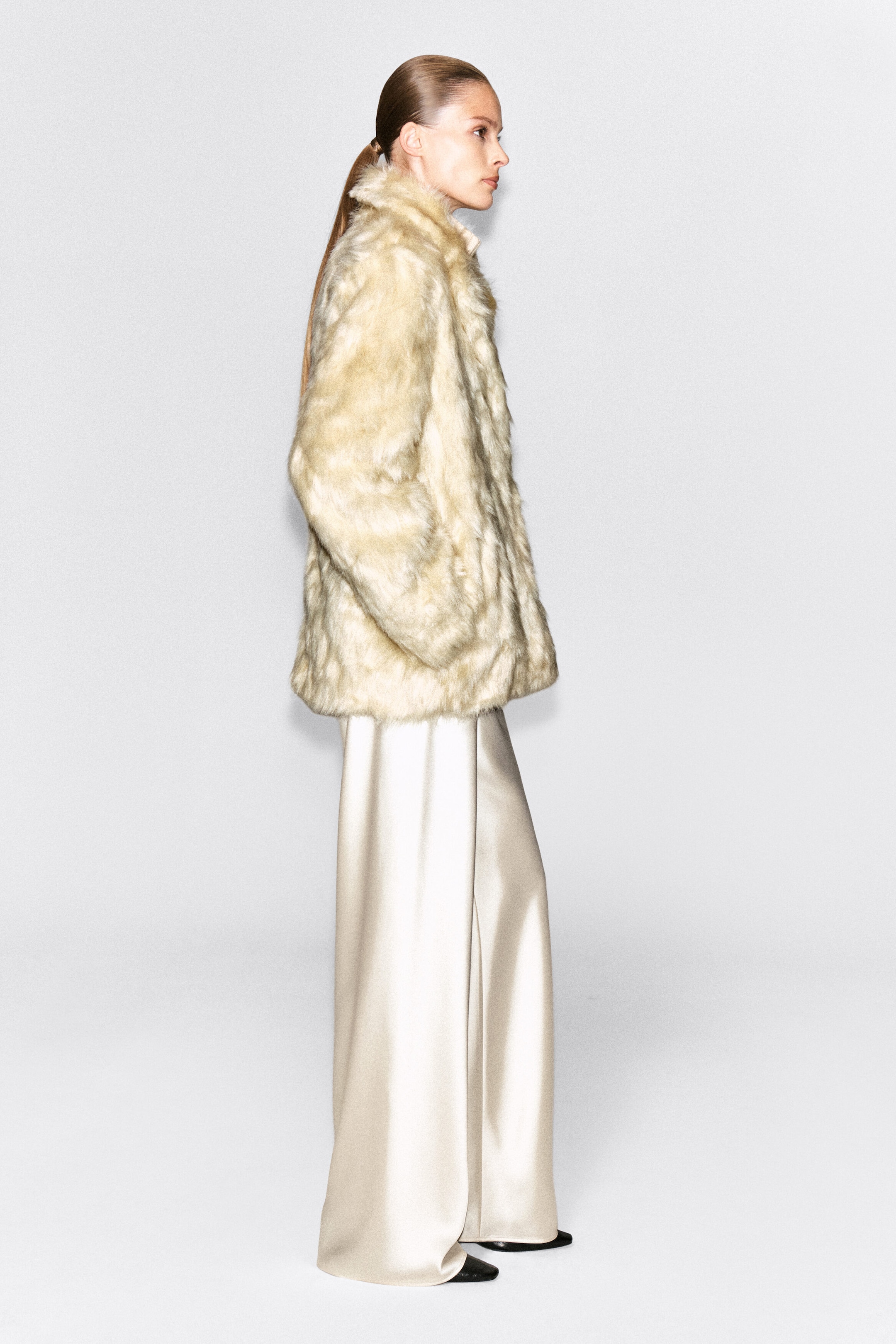 The model is wearing a cream-colored fur-effect jacket in a side-profile pose. The jacket features a plush, textured finish and is styled with wide-leg cream satin trousers.
