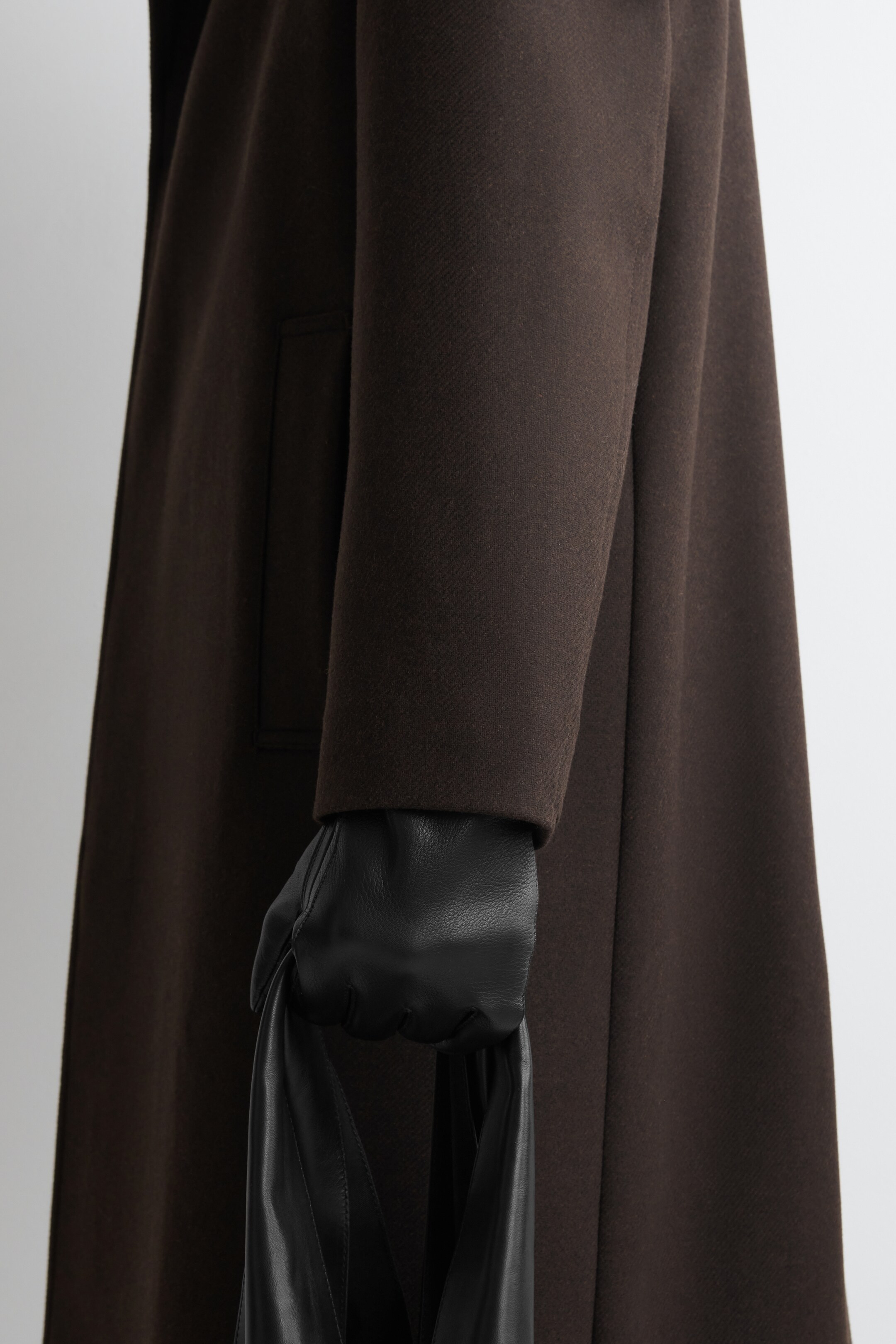 View larger image: A dark brown coat, featuring a smooth, slightly textured fabric and a subtle pocket flap on its side, is being presented from a side angle. A hand, clad in a black leather glove, is emerging from the coat's sleeve and holding a black leather bag handle.