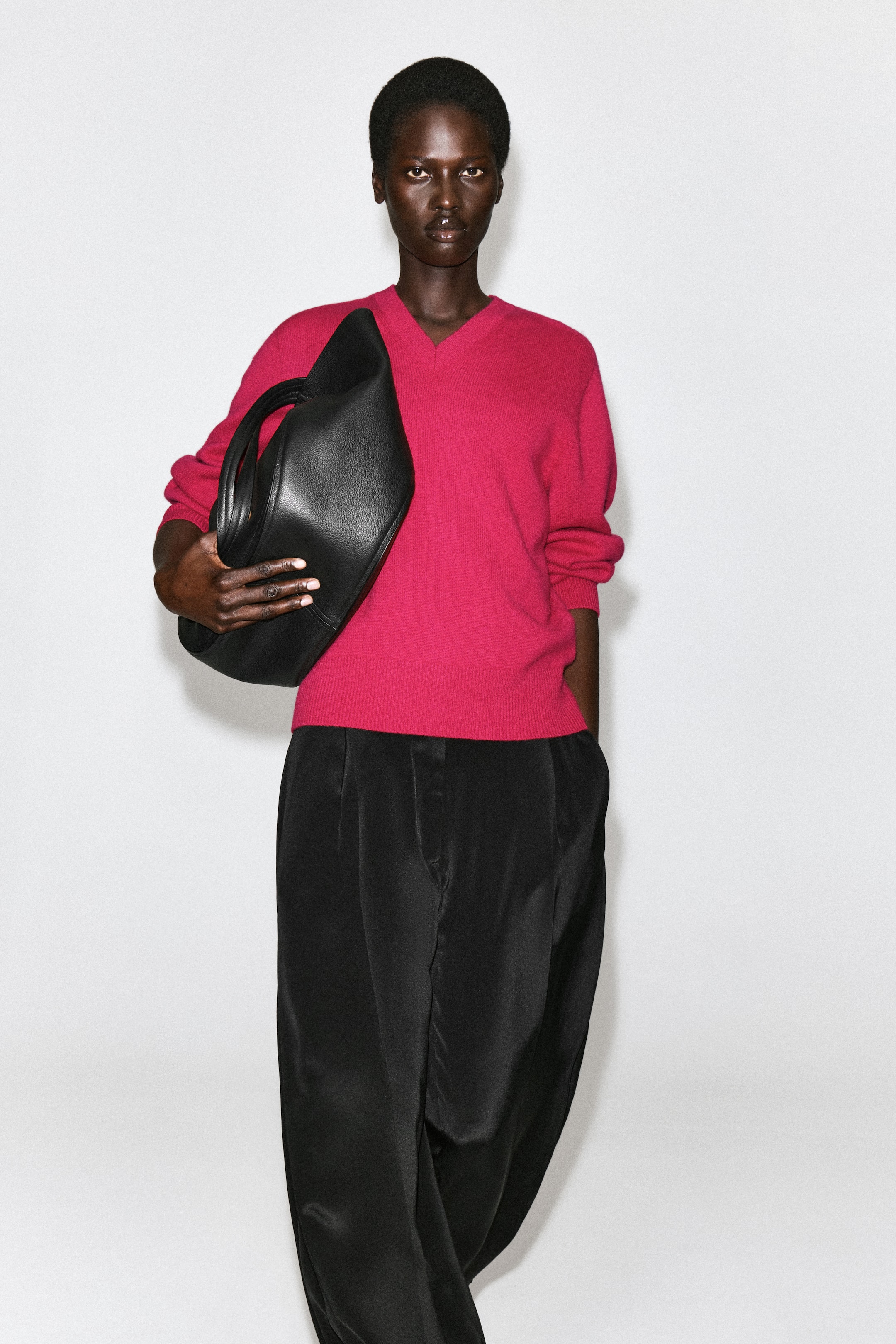 Ver imagen más grande: El jersey de cuello de pico de cachemira pura, de corte estructurado y en un vibrante rosa, lo lleva la modelo, con un clásico escote en V y remates acanalados. El jersey se combina con un pantalón negro, y la modelo lleva un gran bolso de cuero negro bajo un brazo.
