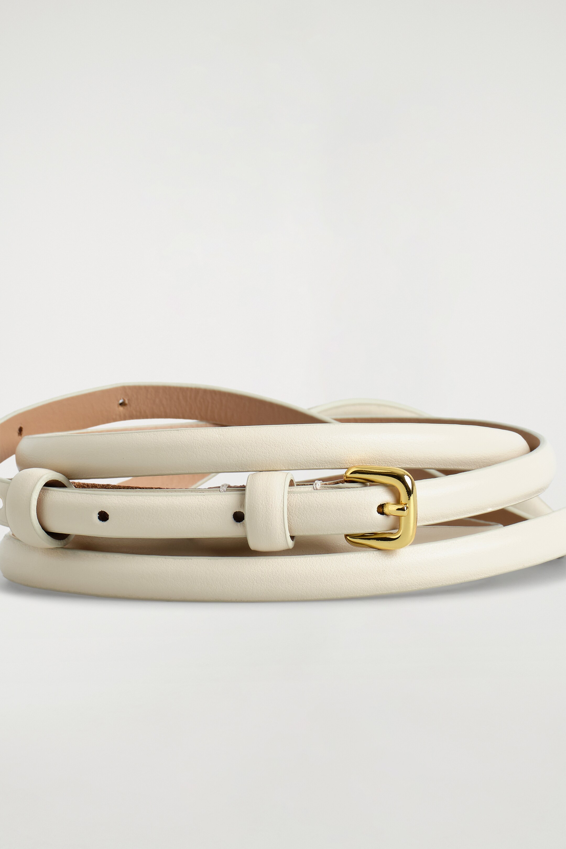 Zobrazit větší obrázek: Úzký, krémově zbarvený kožený pásek se vine, přičemž jeho zlatavá, obdélníková spona s jedním trnem směřuje dopředu.