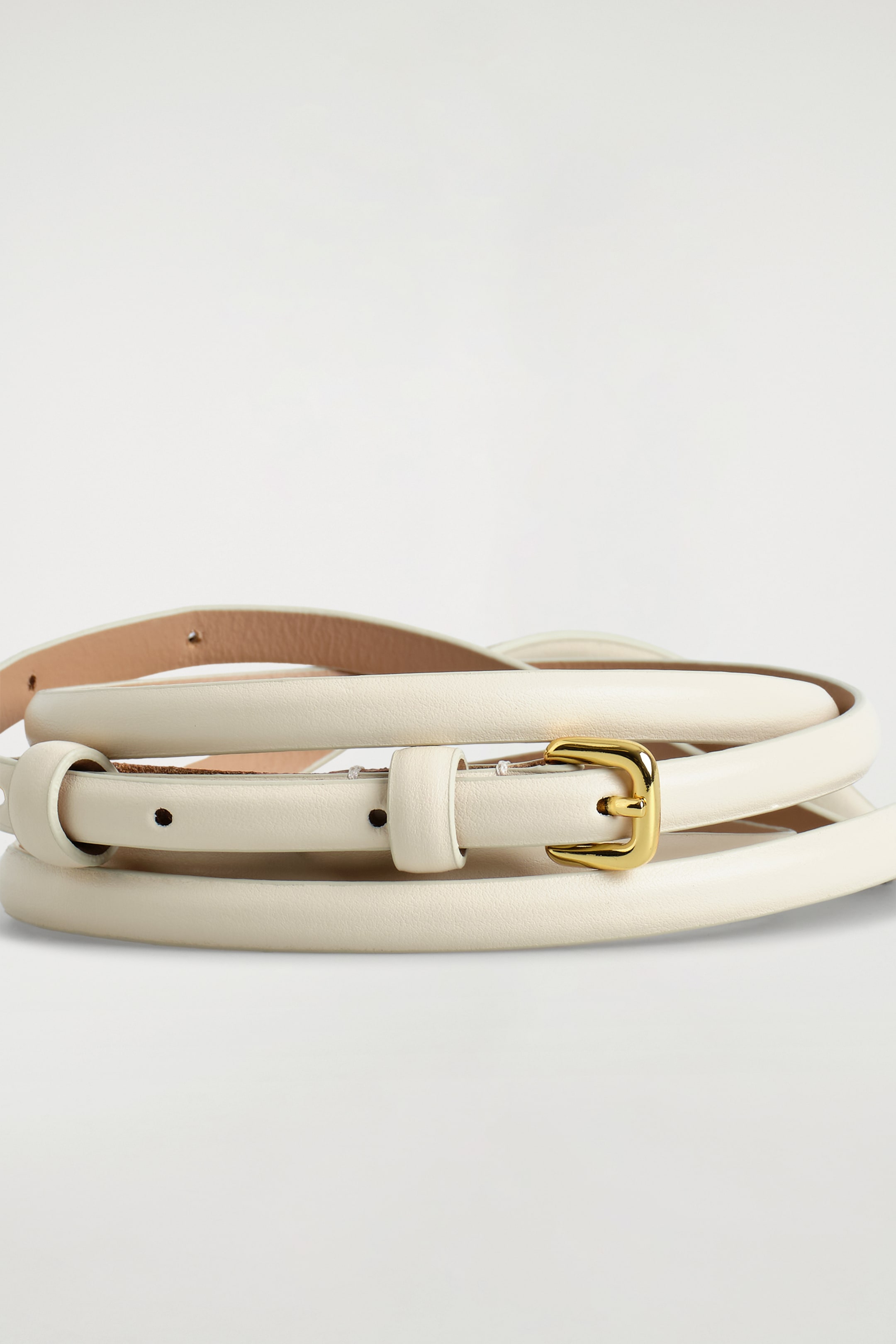 Agrandir l'image: Une ceinture fine en cuir couleur crème s'enroule, avec sa boucle rectangulaire dorée et son ardillon unique orientés vers l'avant.
