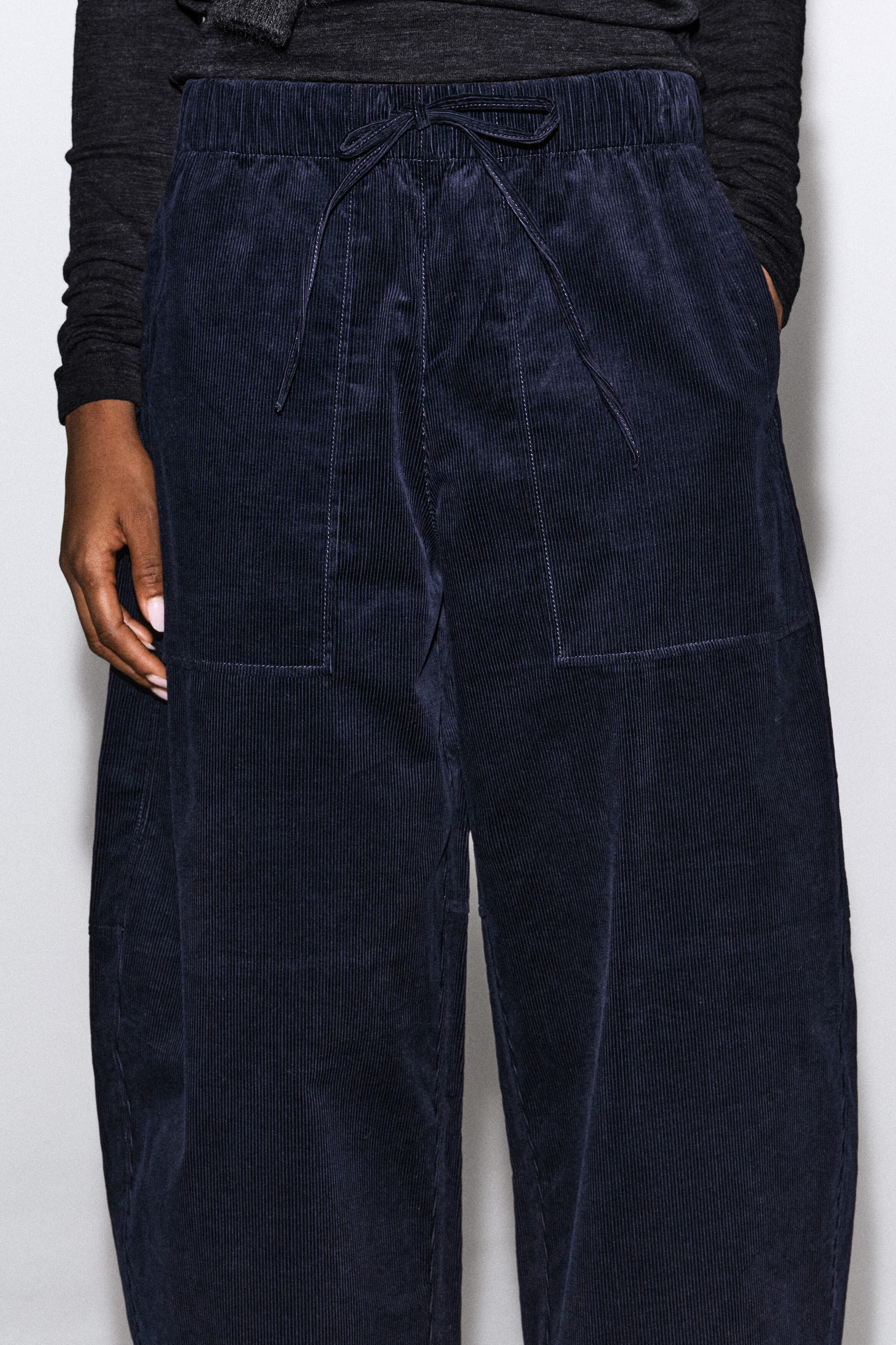 The model is wearing navy blue corduroy barrel-leg drawstring pants, featuring an elasticized waistband with a tied drawstring and prominent front patch pockets. The model is standing with one hand resting in a pocket, showcasing the relaxed fit and fine-wale texture of the fabric. The model is wearing navy blue corduroy barrel-leg drawstring pants, featuring an elasticized waistband with a tied drawstring and prominent front patch pockets. The model is standing with one hand resting in a pocket, showcasing the relaxed fit and fine-wale texture of the fabric.