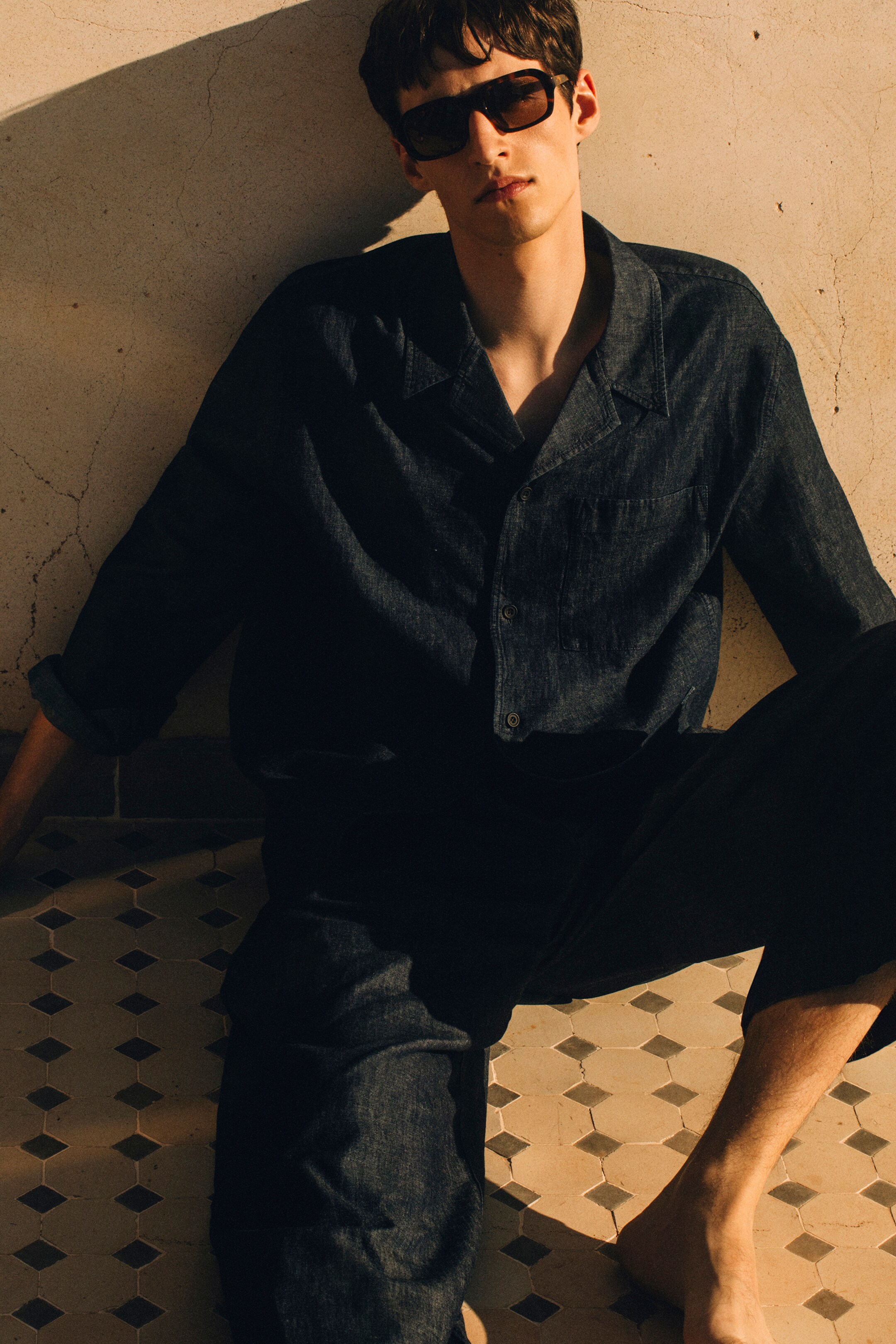 Größeres Bild anzeigen: Ein dunkelblaues Overshirt im Denim-Look mit Resort-Kragen, Knopfleiste vorne und einer einzelnen Brusttasche wird von vorne präsentiert. Es wird mit passenden, locker sitzenden Barrel-Leg-Hosen kombiniert, die leicht gekürzt sind, und die Ärmel des Overshirts sind hochgekrempelt.