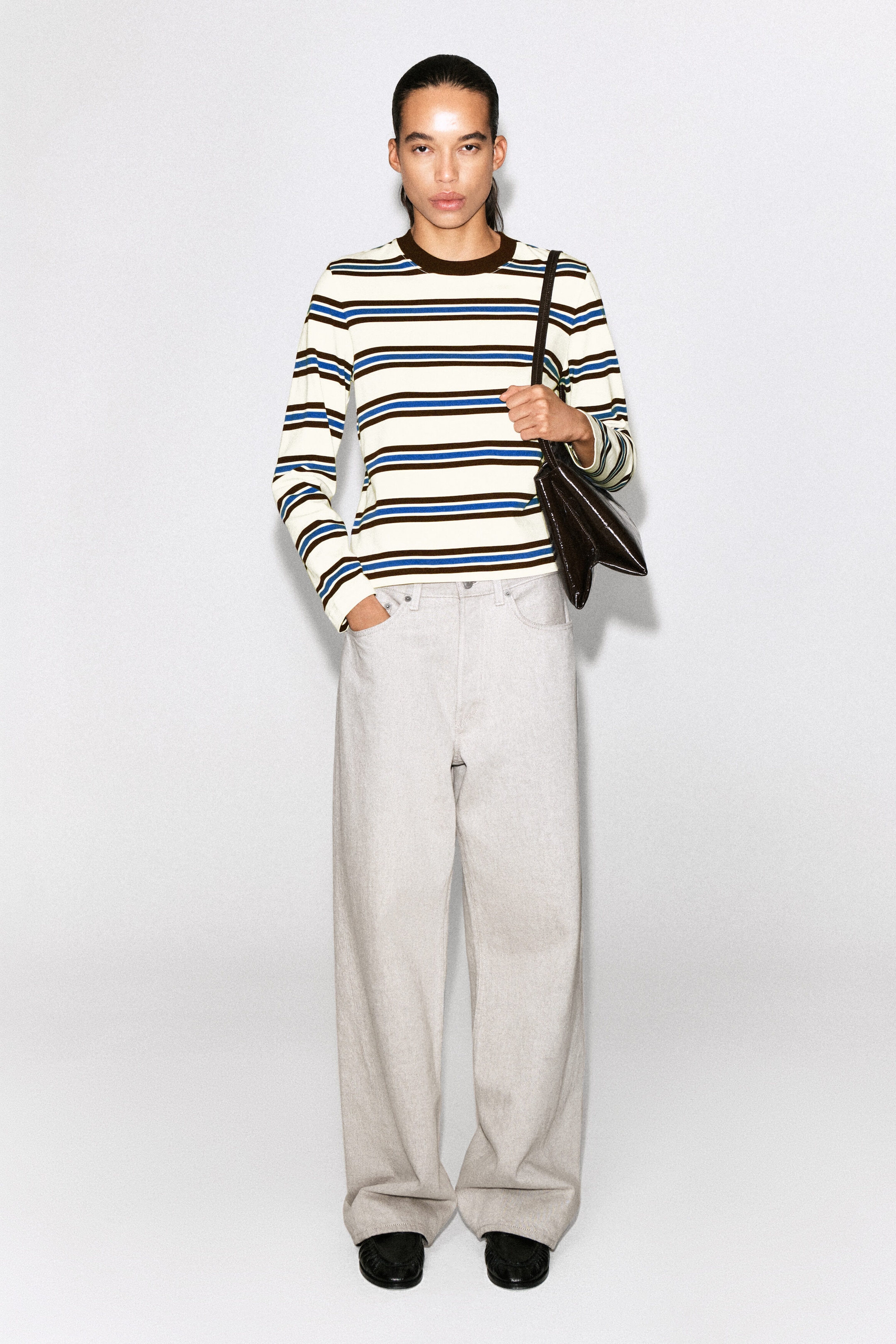 A clean-cut long-sleeved T-shirt features horizontal stripes in off-white, blue and brown, styled front-facing on a model. The T-shirt showcases a contrasting solid brown crew neck and a straight hem, sitting loosely over light-coloured wide-leg trousers. A clean-cut long-sleeved T-shirt features horizontal stripes in off-white, blue and brown, styled front-facing on a model. The T-shirt showcases a contrasting solid brown crew neck and a straight hem, sitting loosely over light-coloured wide-leg trousers.