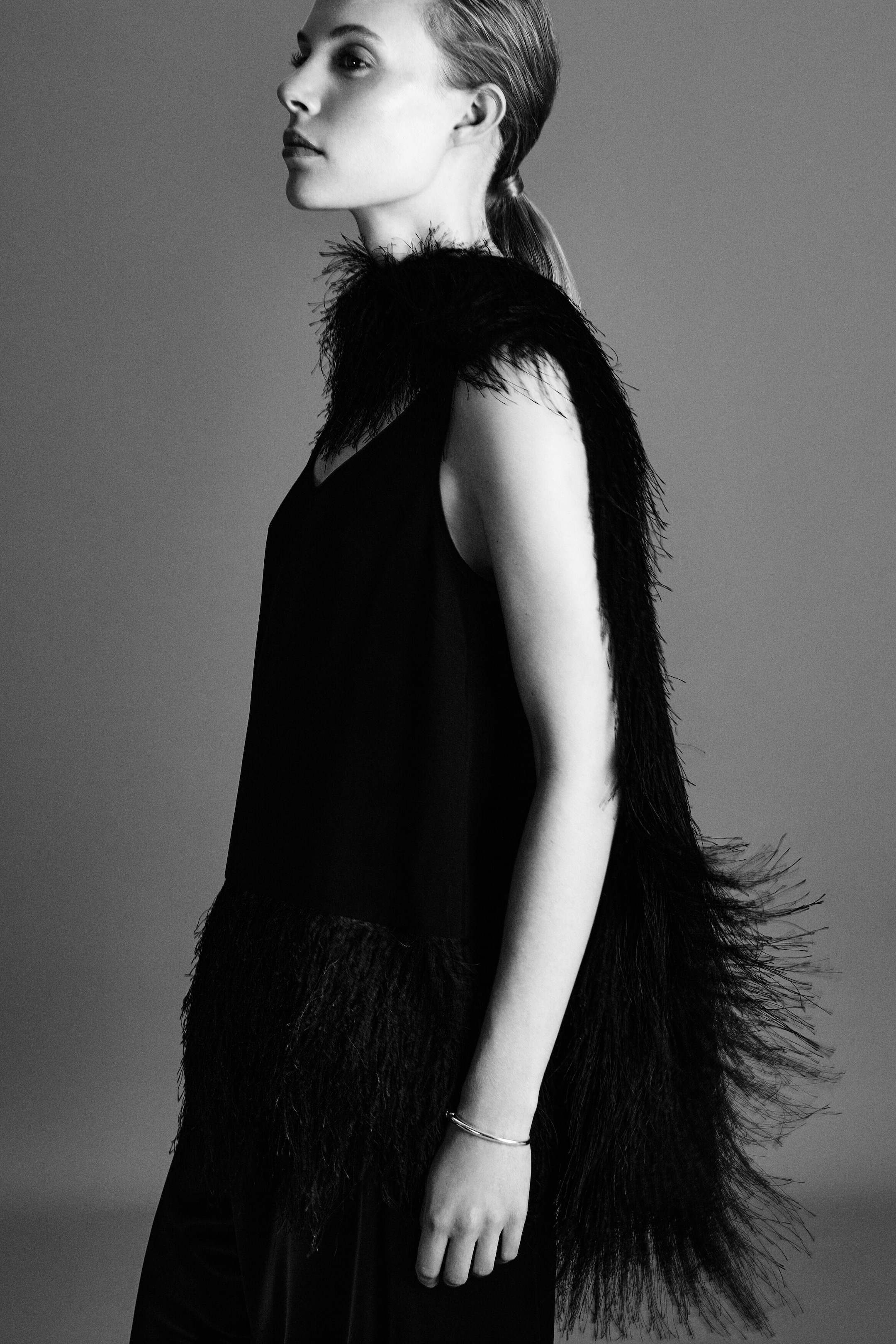A model is standing in side profile, wearing a black sleeveless cami top with a heavily textured, fringed hem, paired with black satin barrel-leg trousers. A long, matching black scarf, adorned with wispy, feathery fringing, is draped over the shoulder and flows down the back to emphasise its movement and texture.