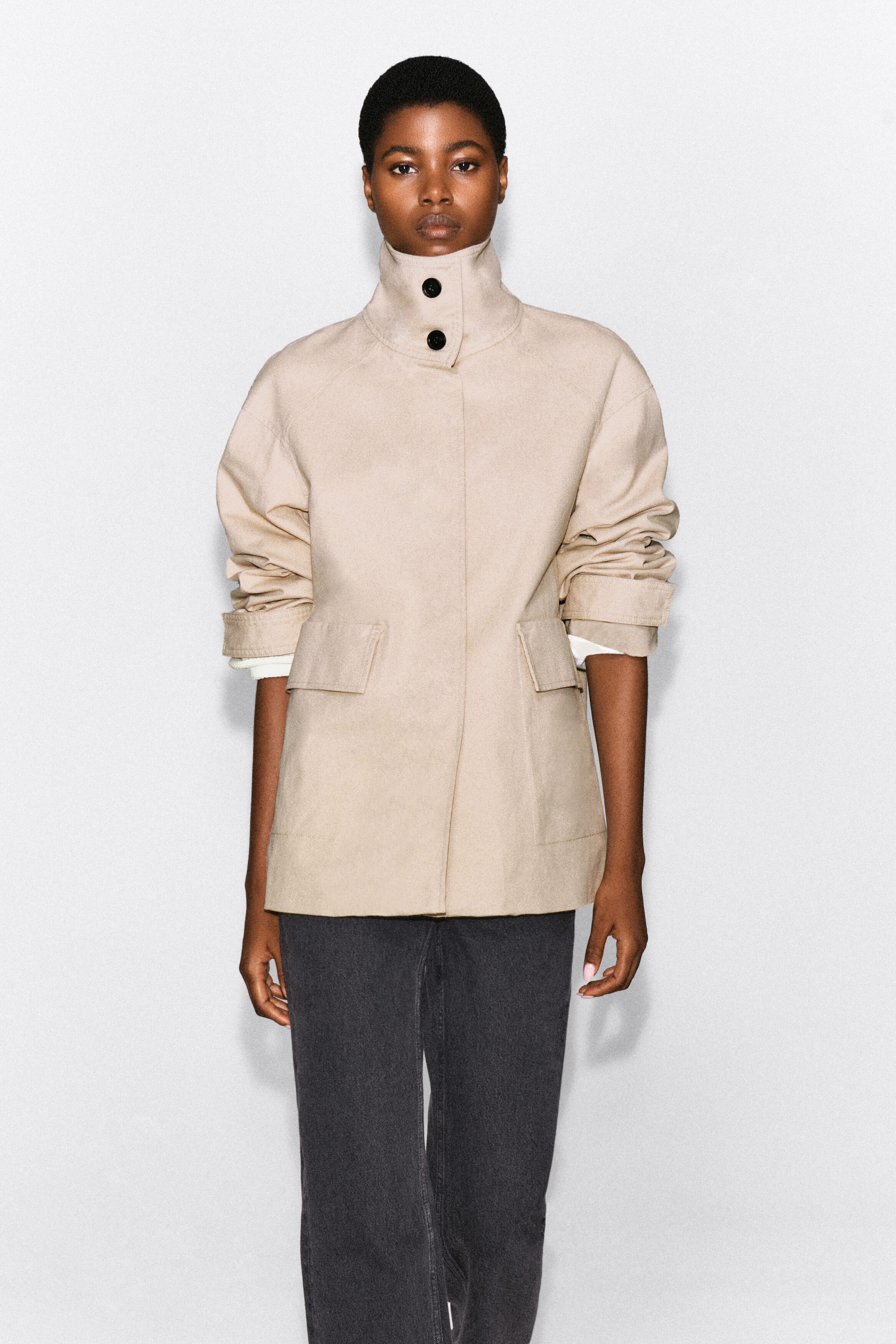 A beige cotton twill funnel-neck jacket is being worn by the model, featuring a high collar with two dark buttons, a concealed front placket, and large flap pockets. The jacket is paired with dark trousers, and the sleeves are slightly pushed up to reveal white cuffs underneath. A beige cotton twill funnel-neck jacket is being worn by the model, featuring a high collar with two dark buttons, a concealed front placket, and large flap pockets. The jacket is paired with dark trousers, and the sleeves are slightly pushed up to reveal white cuffs underneath.