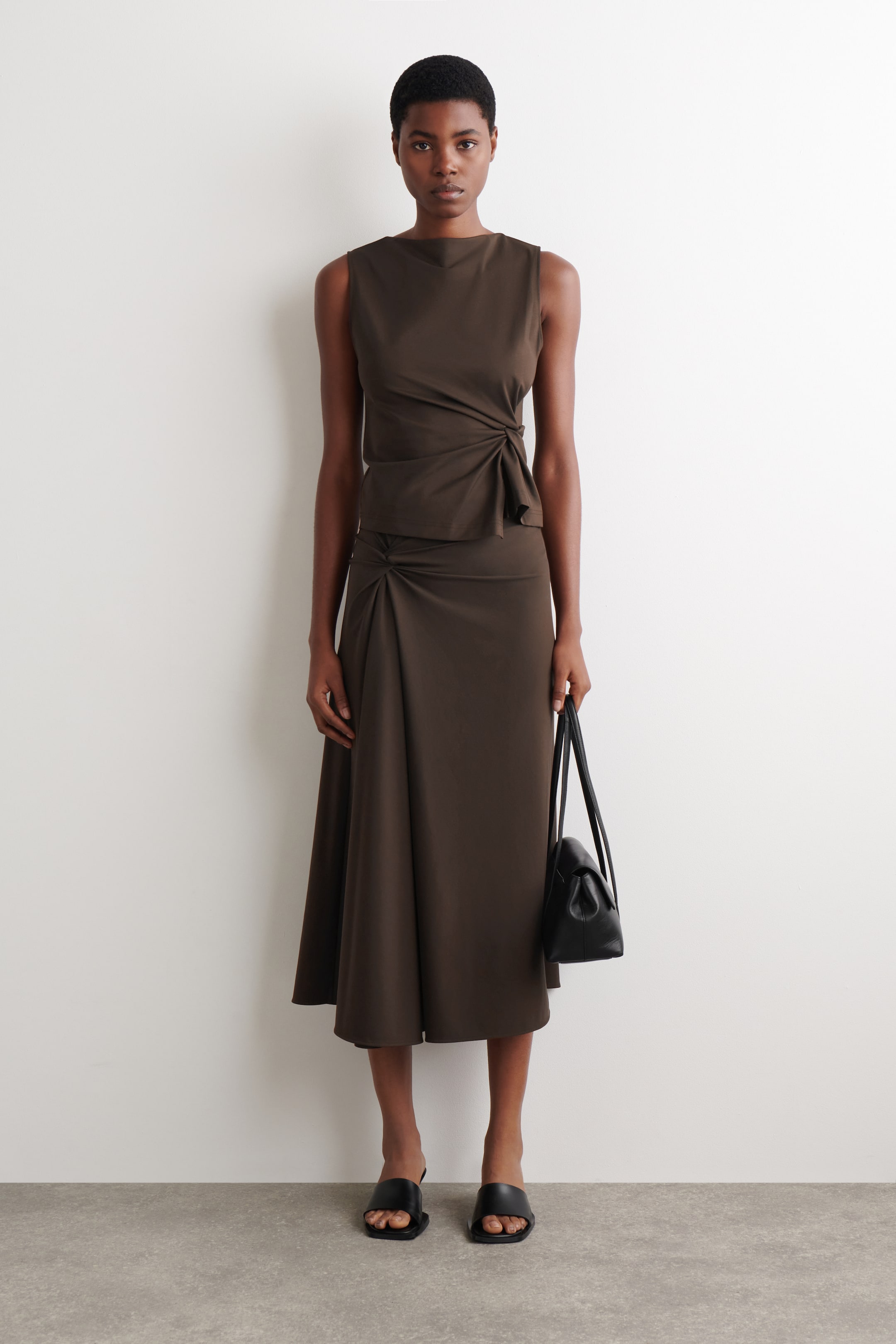 הצגת תמונה גדולה יותר: חולצה ללא שרוולים בצבע זית כהה עם מחשוף בצורת סירה ופרט קשר מקופל במותן השמאלית, משתלבת עם חצאית מידי זורמת שגם היא כוללת פרט קשר מקופל בירך השמאלית.