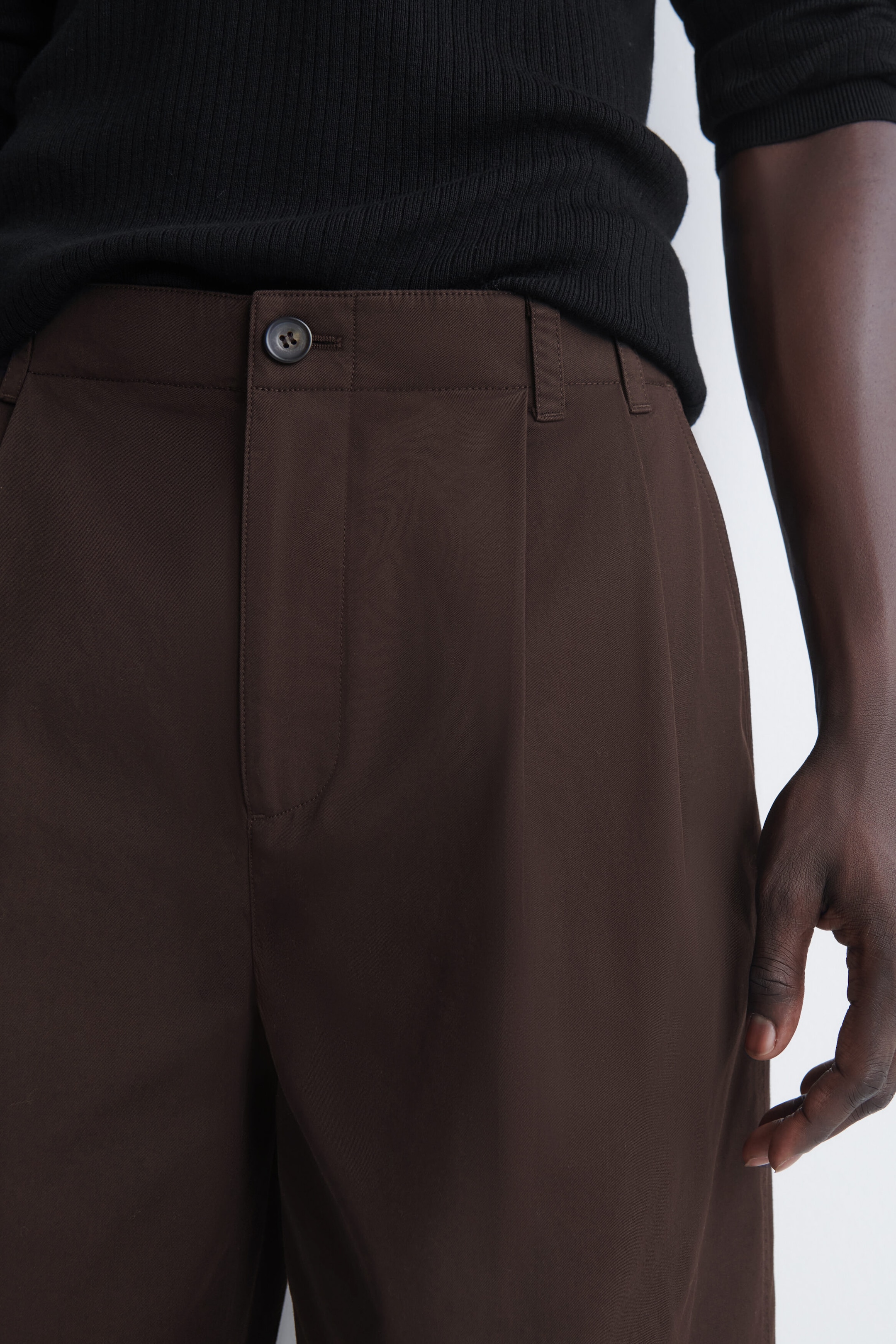The tailored pleated cotton shorts in dark brown are facing front, showcasing a structured waistband with belt loops, a single button fastening, and a sharp front pleat. A black ribbed top is tucked into the waistband, highlighting the clean, tailored fit of the shorts. The tailored pleated cotton shorts in dark brown are facing front, showcasing a structured waistband with belt loops, a single button fastening, and a sharp front pleat. A black ribbed top is tucked into the waistband, highlighting the clean, tailored fit of the shorts.