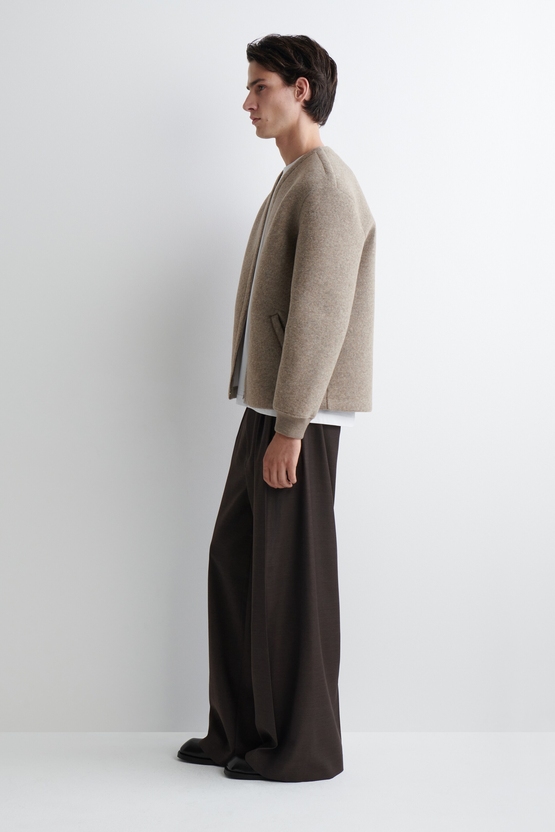 Agrandir l'image: Une veste marron clair sans col, avec des épaules tombantes et des poches passepoilées à l'avant, est portée par-dessus un t-shirt blanc et associée à un pantalon marron foncé à jambes larges qui s'étend sur des chaussures noires. L'ensemble est présenté de côté, mettant en valeur la coupe décontractée de la veste et le drapé fluide du pantalon.