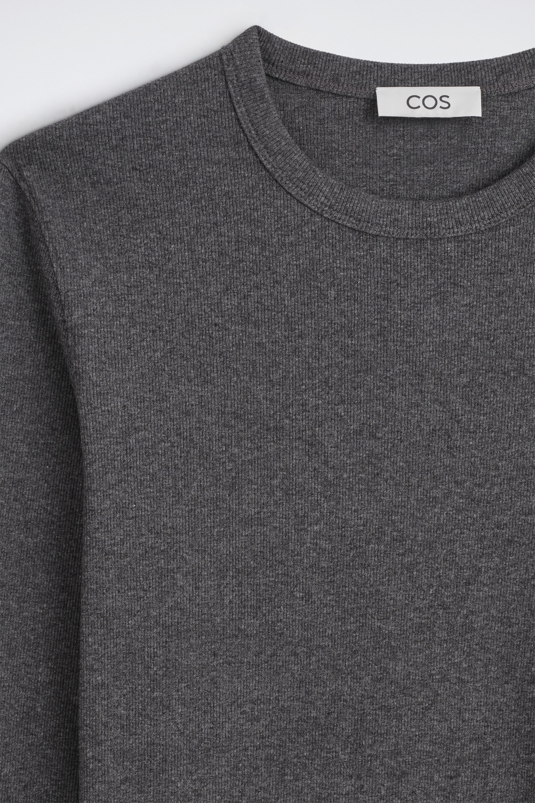 A dark grey ribbed knit top is displayed in a close-up front view, featuring a classic crew neckline with a ribbed band. The fabric showcases a fine vertical ribbed texture, with a white rectangular brand label stitched inside the back collar. A dark grey ribbed knit top is displayed in a close-up front view, featuring a classic crew neckline with a ribbed band. The fabric showcases a fine vertical ribbed texture, with a white rectangular brand label stitched inside the back collar.