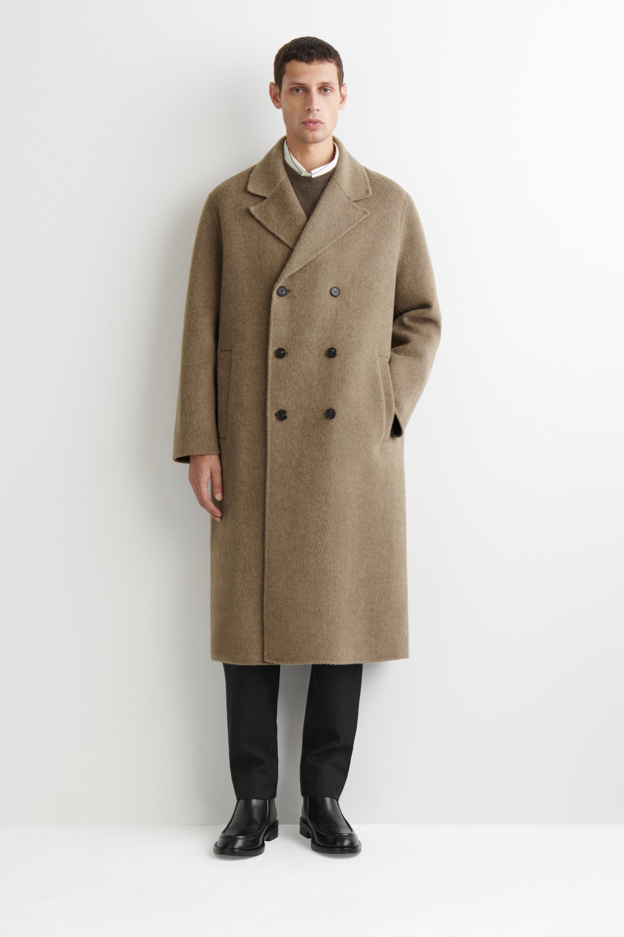 The double-faced wool-blend long coat is shown from the front, highlighting a relaxed double-breasted silhouette with wide notch lapels and a six-button closure. The textured beige-brown coat is layered over a brown sweater and white collared shirt, falling below the knee and paired with black trousers and leather boots.