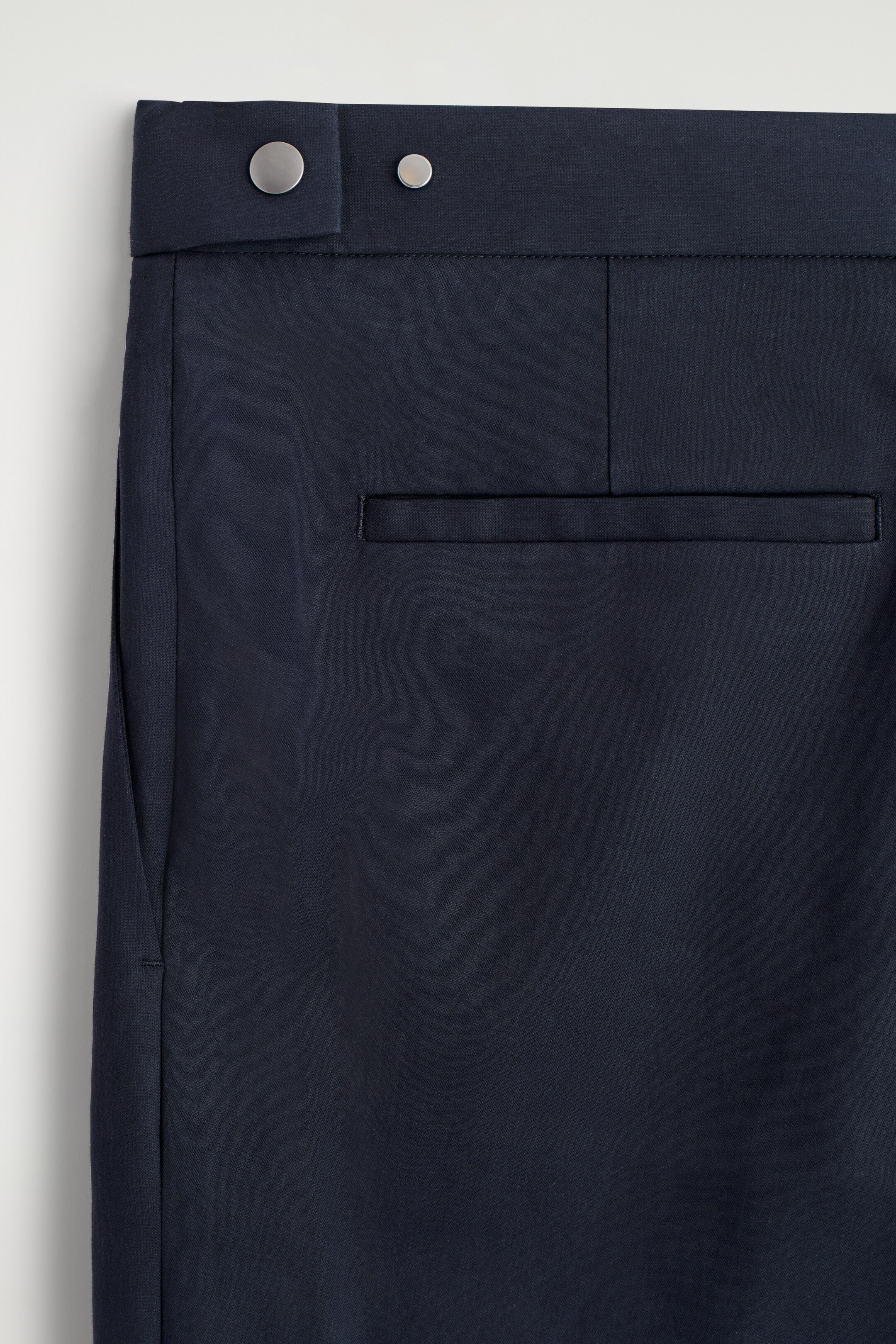 View larger image: A close-up of the back of black slim trousers is displaying a smooth, dark cotton fabric. The streamlined waistband is featuring a large silver-toned press stud on an extended tab and a smaller silver-toned stud, while a single horizontal jetted pocket is visible below the waistband.