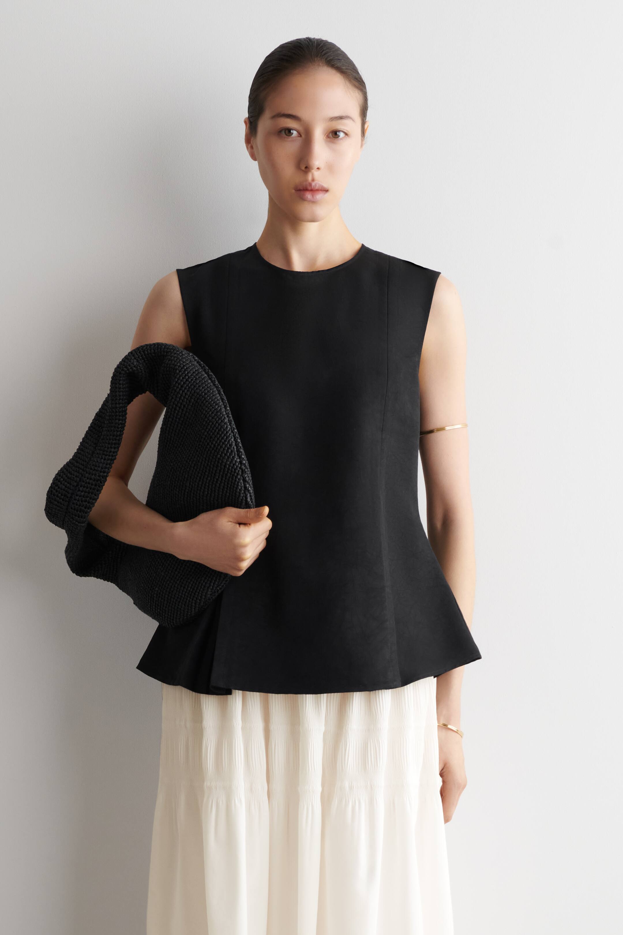 Ver imagen más grande: La modelo lleva puesto un top negro sin mangas con paneles pespunteados y un bajo peplum con doble pliegue, combinado con una falda plisada de color claro.