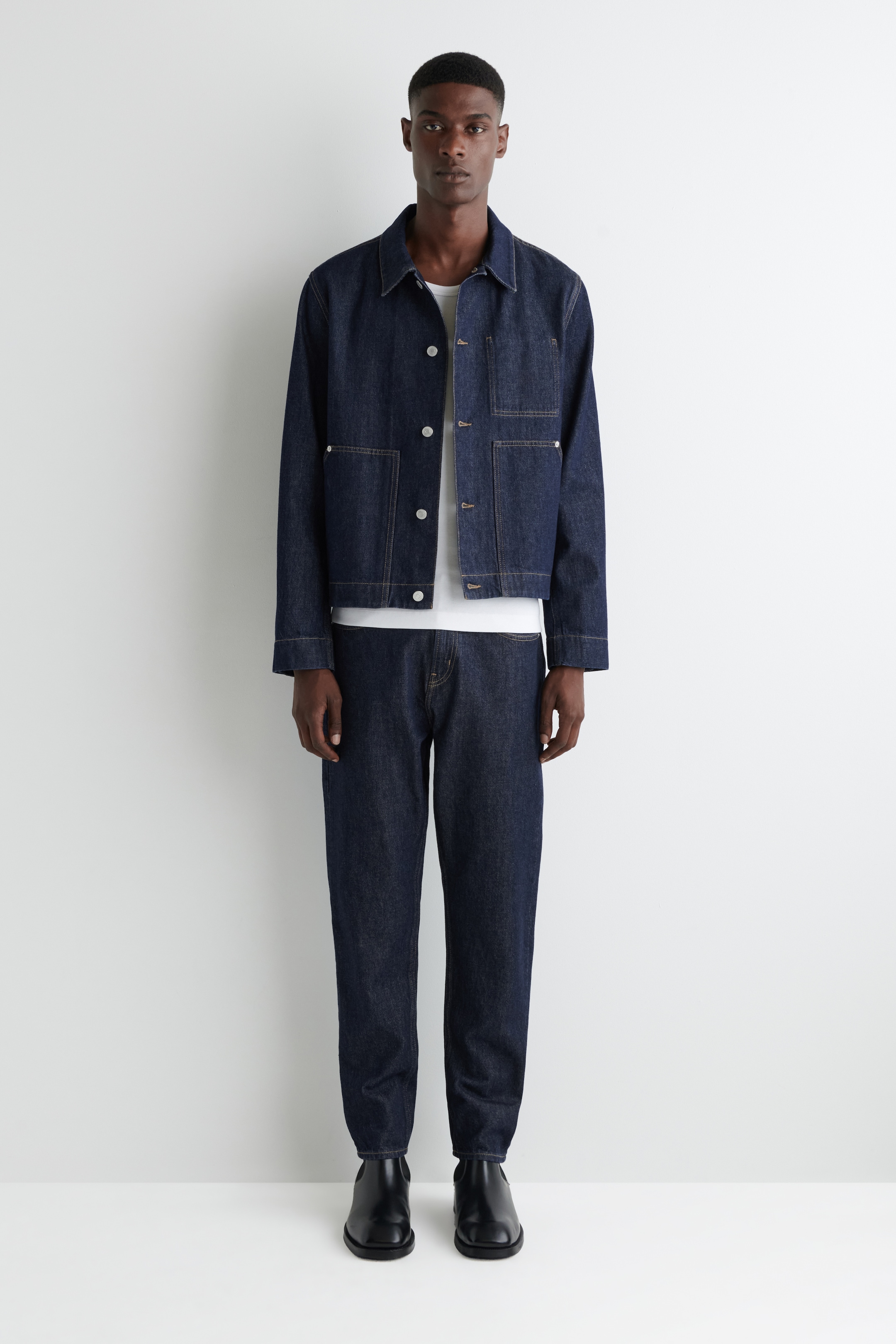 Visualizza immagine più grande: Un abito in denim indaco scuro, composto da una giacca e pantaloni coordinati, è presentato su una persona rivolta in avanti. La giacca presenta un colletto a punta, una chiusura frontale con bottoni bianchi e quattro grandi tasche applicate. Sia la giacca che i pantaloni a vestibilità rilassata sono accentuati con cuciture a contrasto, con la giacca sovrapposta a una t-shirt bianca e i pantaloni abbinati a stivali neri.