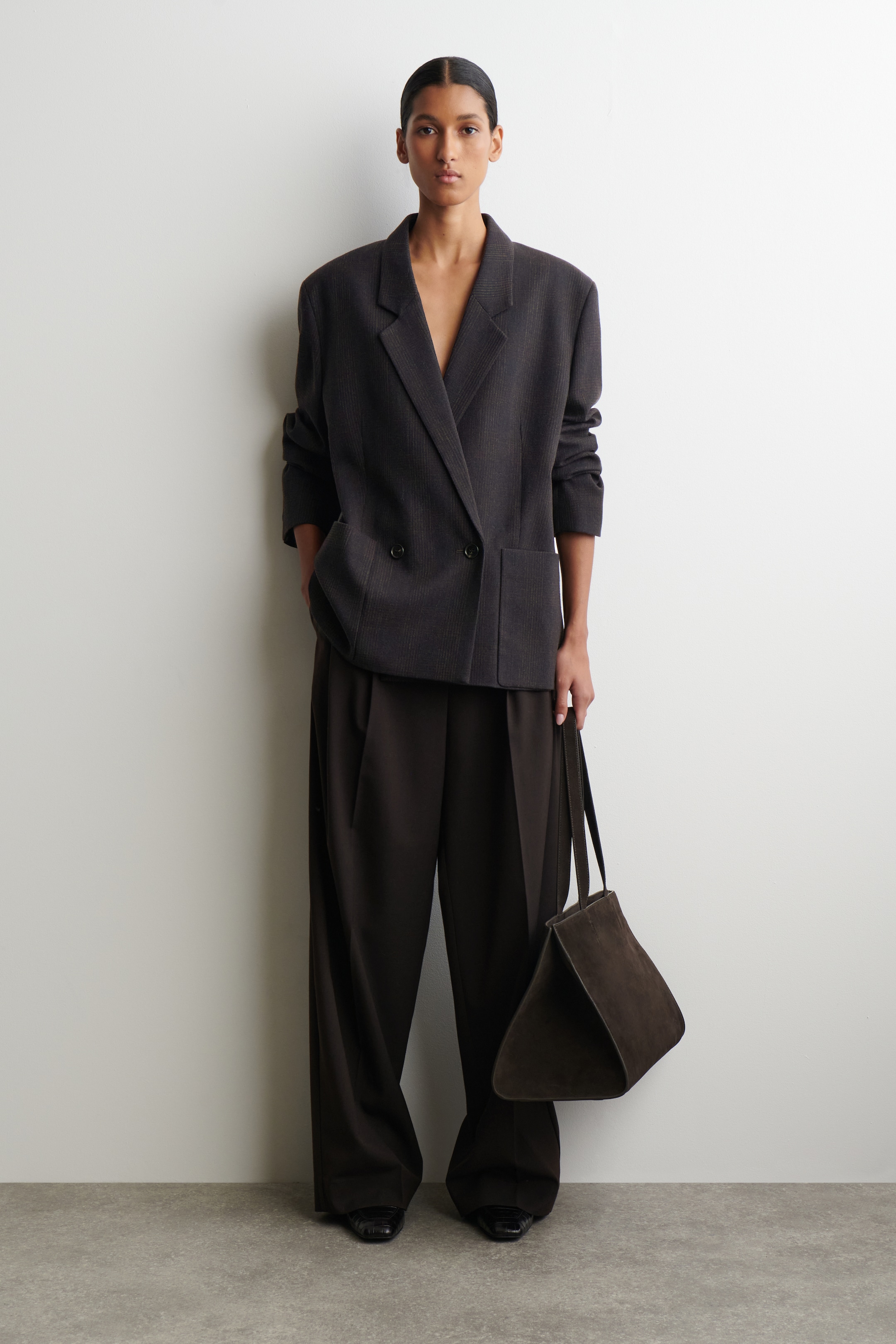 Visualizza immagine più grande: Un blazer doppiopetto a righe sottili grigio scuro viene presentato in una visuale frontale completa, caratterizzato da una vestibilità oversize, revers a dente e tasche con pattina visibili. È abbinato a pantaloni a gamba larga con pieghe di colore marrone scuro, e una borsa a secchiello geometrica di camoscio marrone scuro viene tenuta al lato.