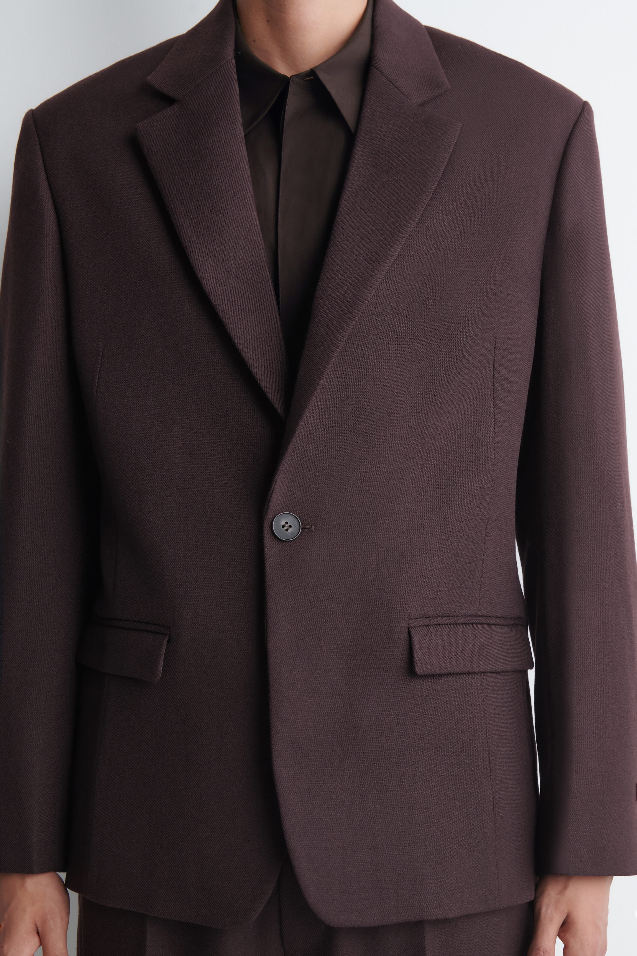 The model is wearing a dark brown textured blazer featuring notch lapels, a single-button fastening, and two flap pockets. A dark brown collared shirt is visible underneath. The model is wearing a dark brown textured blazer featuring notch lapels, a single-button fastening, and two flap pockets. A dark brown collared shirt is visible underneath.