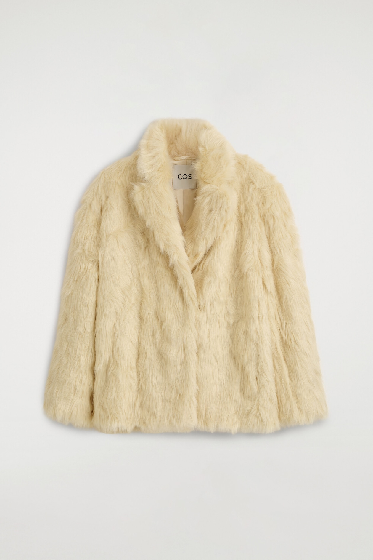 A cream-colored, fur-effect jacket is shown from the front, highlighting its plush, textured surface and relaxed silhouette. The design features wide notch lapels, a wrap-front closure, and long, straight sleeves.