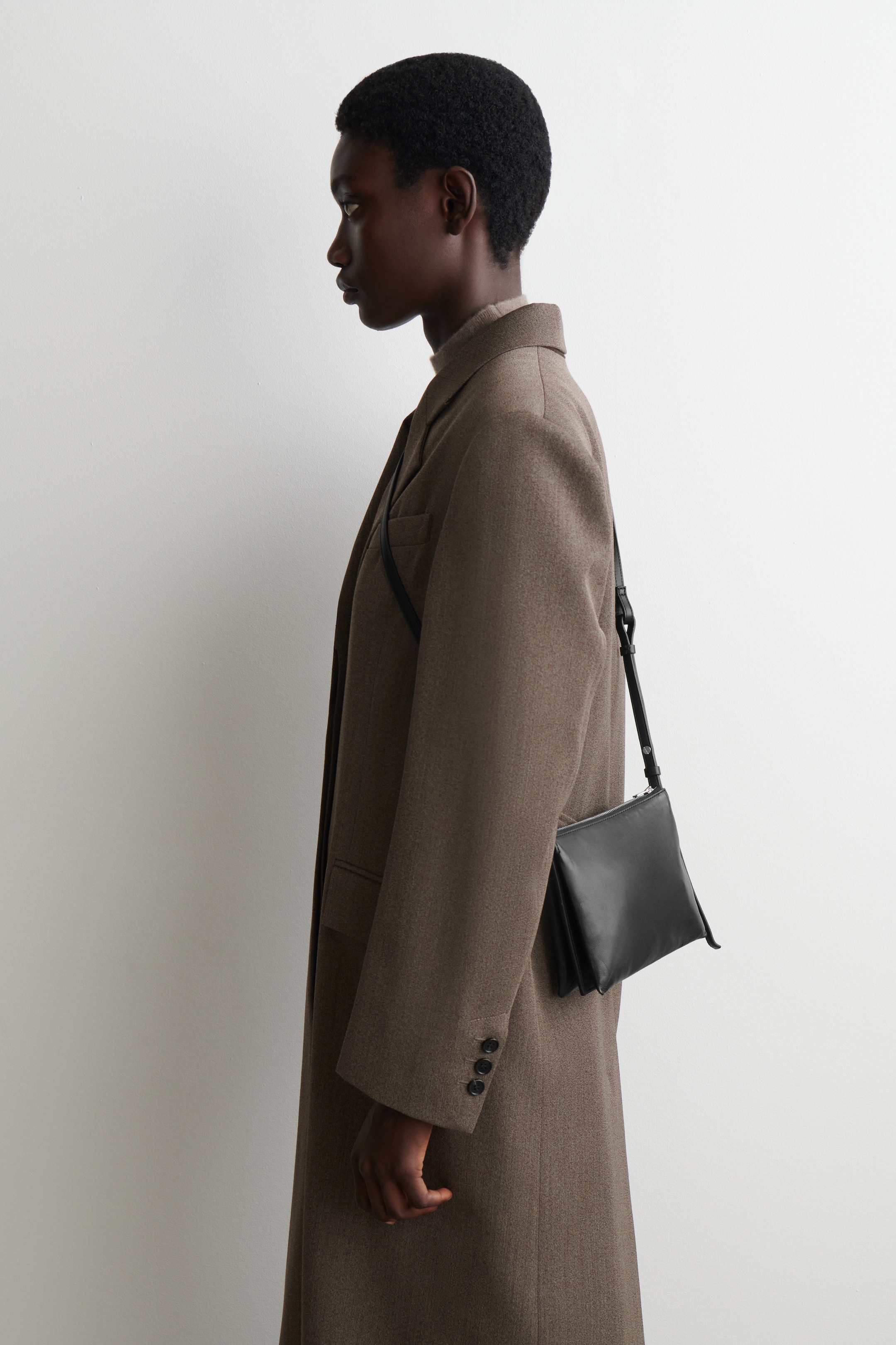 View larger image: A long, earthy brown overcoat is being presented from a side profile, showcasing its straight cut and textured wool-blend fabric. It is featuring notch lapels, a visible flap pocket, and three buttons on the cuff of its long sleeve. A black crossbody bag is being worn over the coat, and a light high-neck top is layered underneath.