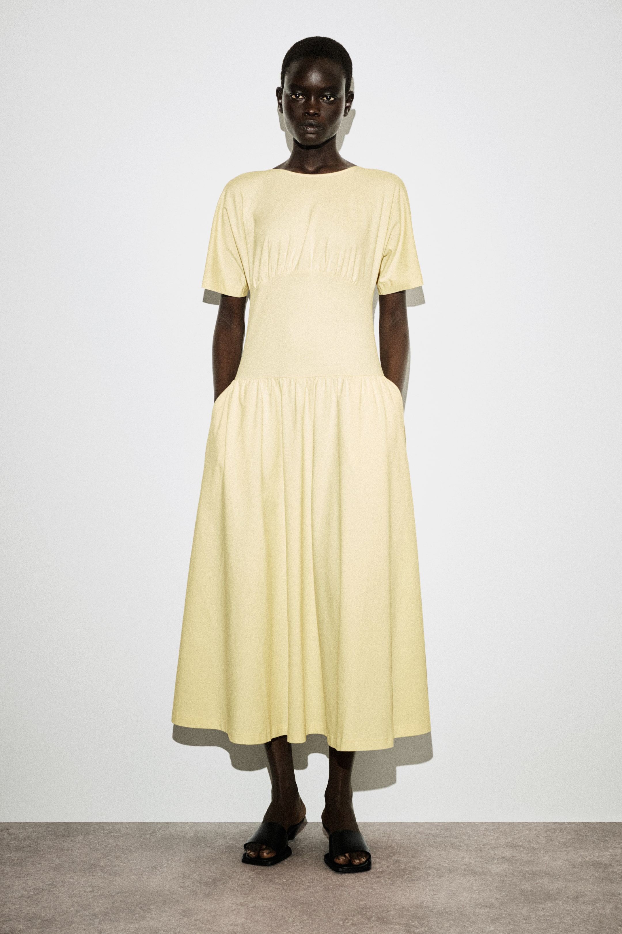 הצגת תמונה גדולה יותר: שמלת מידי בצבע צהוב חיוור פונה קדימה, עם צווארון סירה, שרוולים קצרים וריפוד בחזה. היא מציגה קו מותן אמפיר מוגדר עם חצאית מידי מתנפנפת ומקובצת, שעוצבה עם כיסים בצדדים.