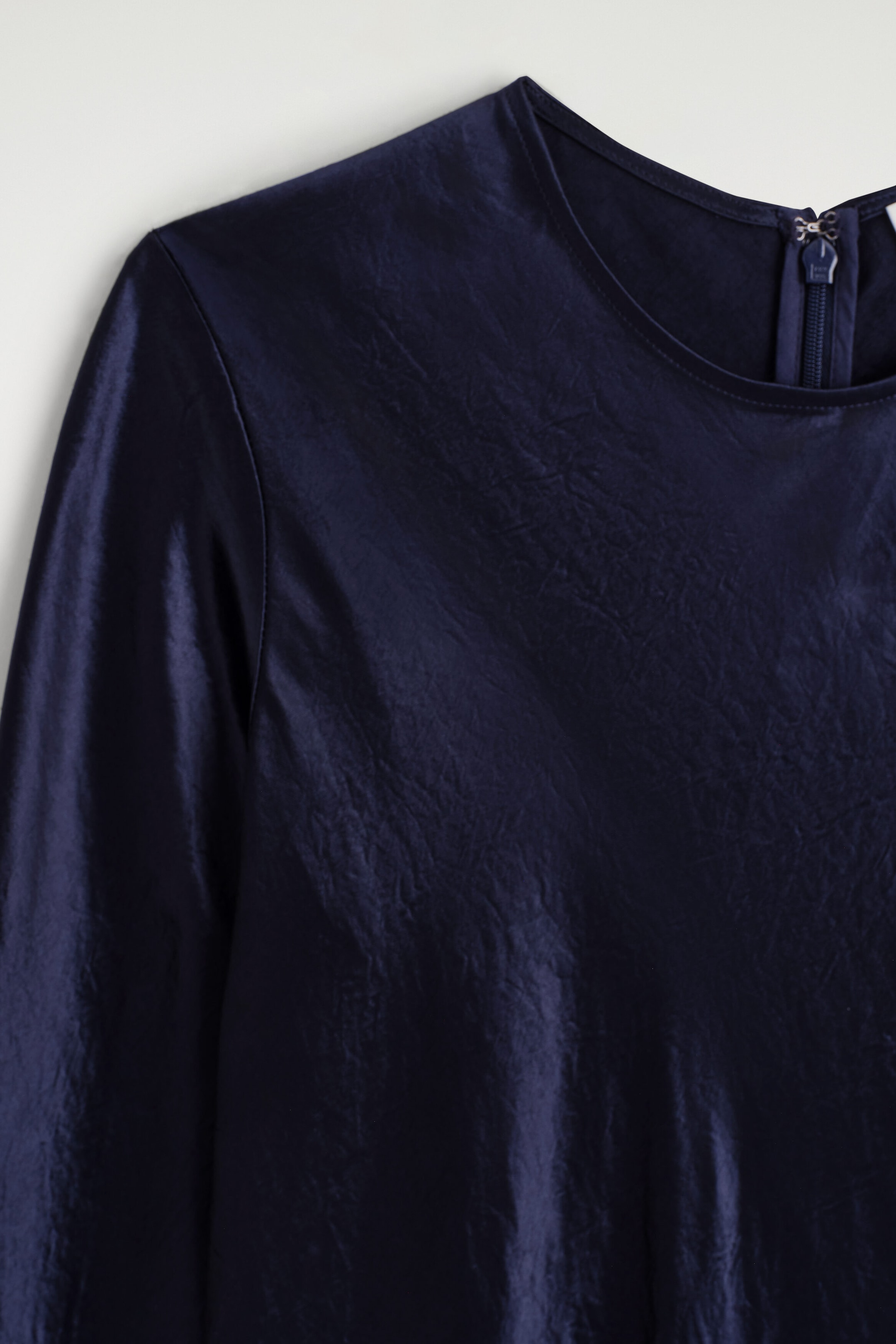 A navy blue satin long-sleeve maxi dress is shown in a close-up view, highlighting its textured fabric with a subtle sheen. The dress features a round neckline, long sleeves, and a partially visible zipper closure at the back of the neck. A navy blue satin long-sleeve maxi dress is shown in a close-up view, highlighting its textured fabric with a subtle sheen. The dress features a round neckline, long sleeves, and a partially visible zipper closure at the back of the neck.