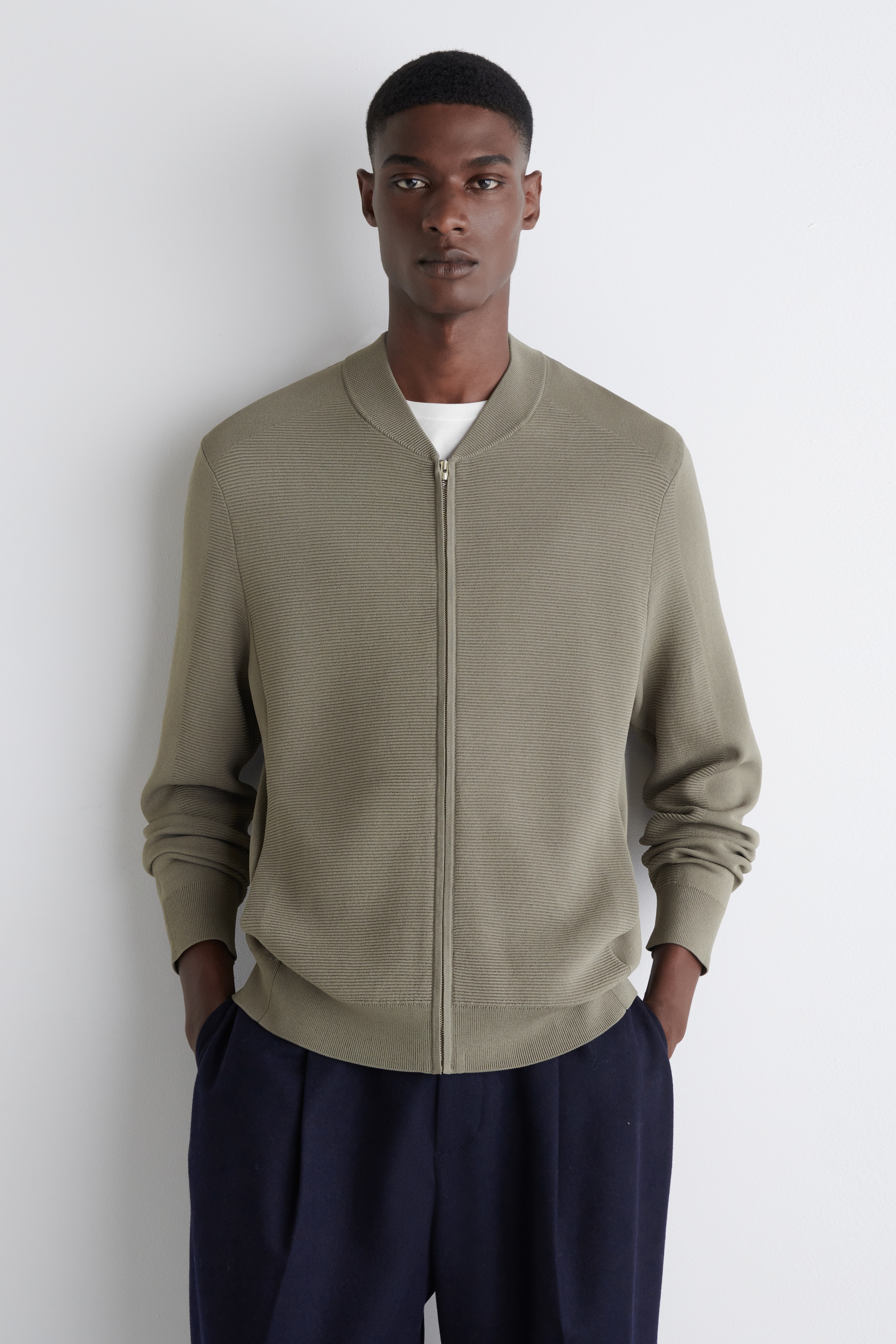 Agrandir l'image: Une veste bomber en maille côtelée vert olive est présentée de face, avec une fermeture éclair pleine longueur, un col montant côtelé, des poignets côtelés et un ourlet côtelé, portée par-dessus un t-shirt blanc.