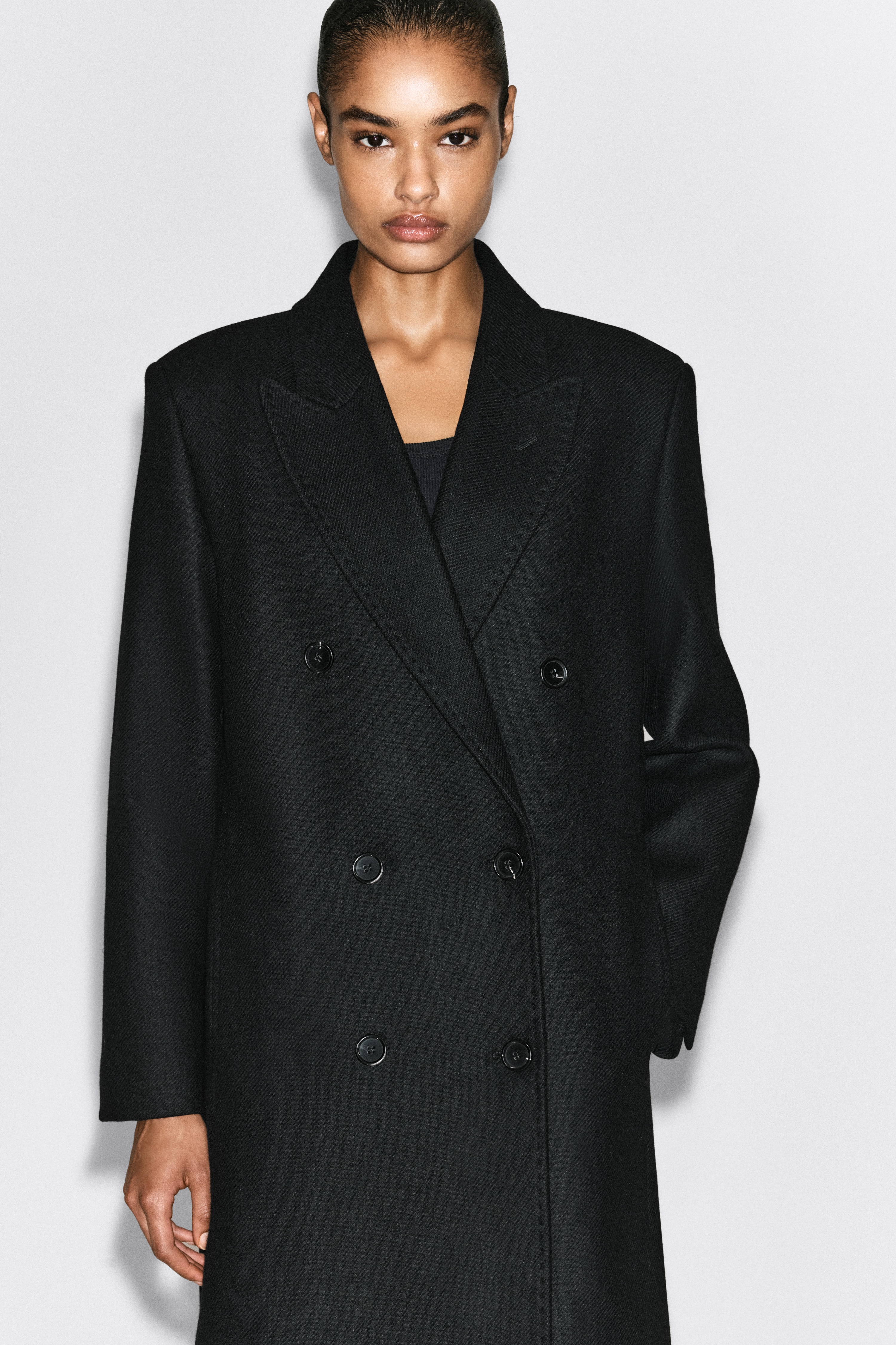 Un manteau noir croisÃ© en mÃ©lange de laine, Ã  la coupe ajustÃ©e, est suspendu de face, mettant en valeur des Ã©paules structurÃ©es, de larges revers crantÃ©s Ã  pointe et une fermeture frontale Ã  six boutons. Ce manteau Ã  manches longues prÃ©sente une silhouette droite mi-longue avec de discrÃ¨tes poches passepoilÃ©es sur les cÃ´tÃ©s.