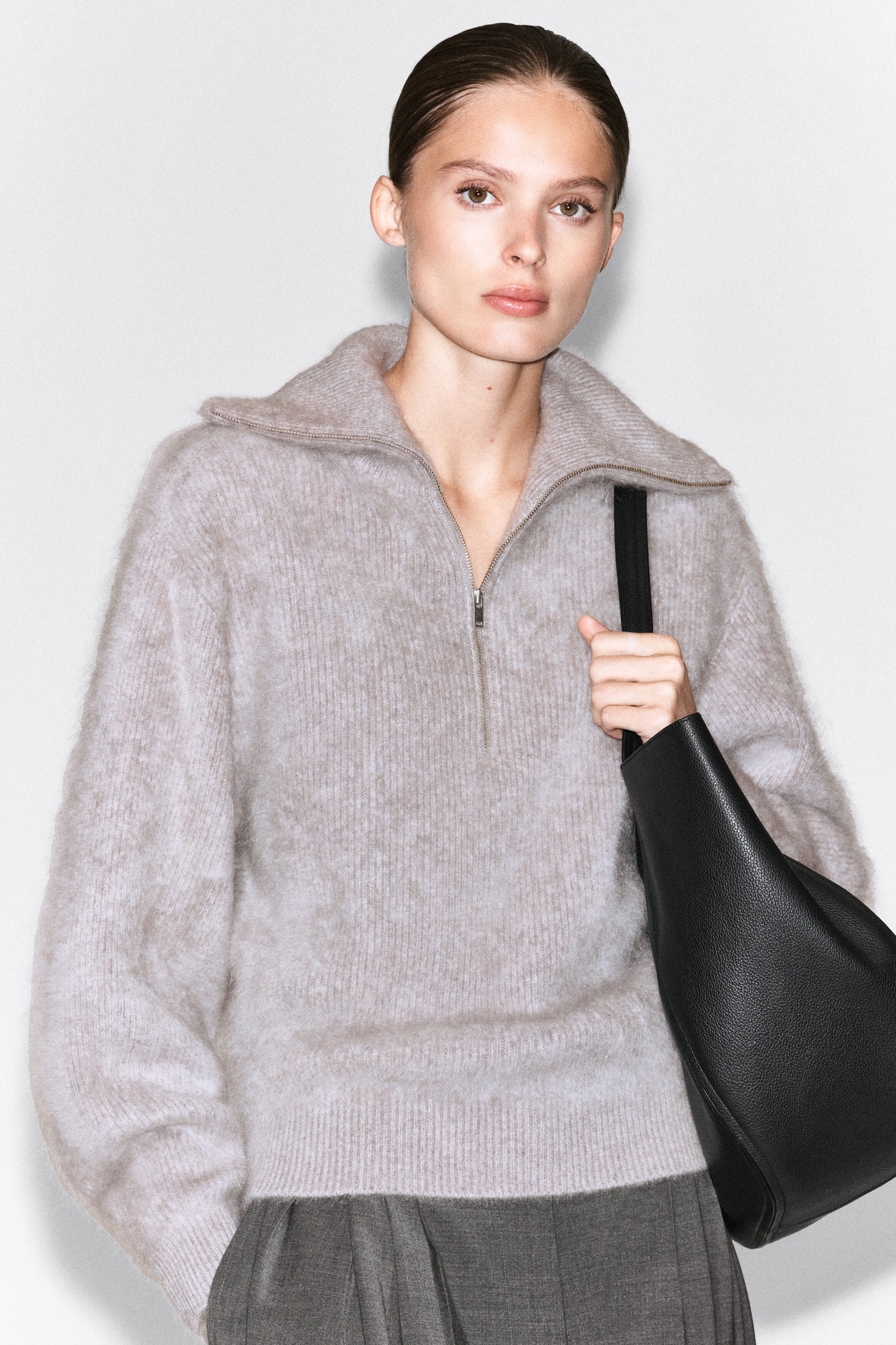 A light gray, brushed cashmere sweater is worn by the model, featuring a silver-tone half-zip at the open funnel collar, ribbed cuffs, and a ribbed hem. The sweater is styled with gray trousers, and the model is holding the strap of a black leather shoulder bag. A light gray, brushed cashmere sweater is worn by the model, featuring a silver-tone half-zip at the open funnel collar, ribbed cuffs, and a ribbed hem. The sweater is styled with gray trousers, and the model is holding the strap of a black leather shoulder bag.