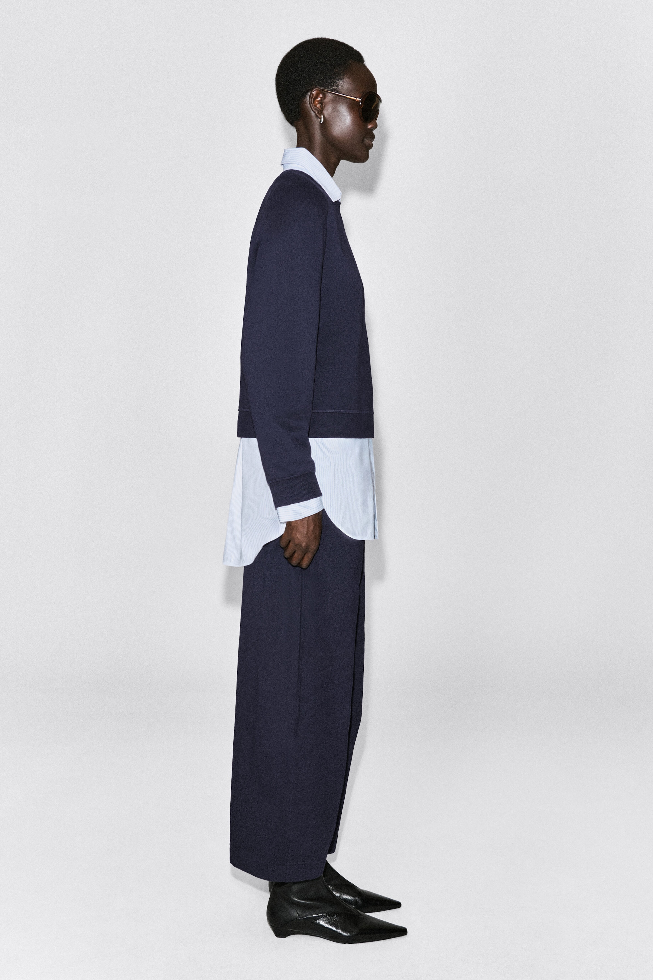 A navy cotton sweatshirt is shown in profile facing right, featuring a crew neck, ribbed cuffs, and a ribbed hem. The sweatshirt is layered over a light-coloured, striped collared shirt that extends past the hem and cuffs, demonstrating its relaxed, structured fit. A navy cotton sweatshirt is shown in profile facing right, featuring a crew neck, ribbed cuffs, and a ribbed hem. The sweatshirt is layered over a light-coloured, striped collared shirt that extends past the hem and cuffs, demonstrating its relaxed, structured fit.