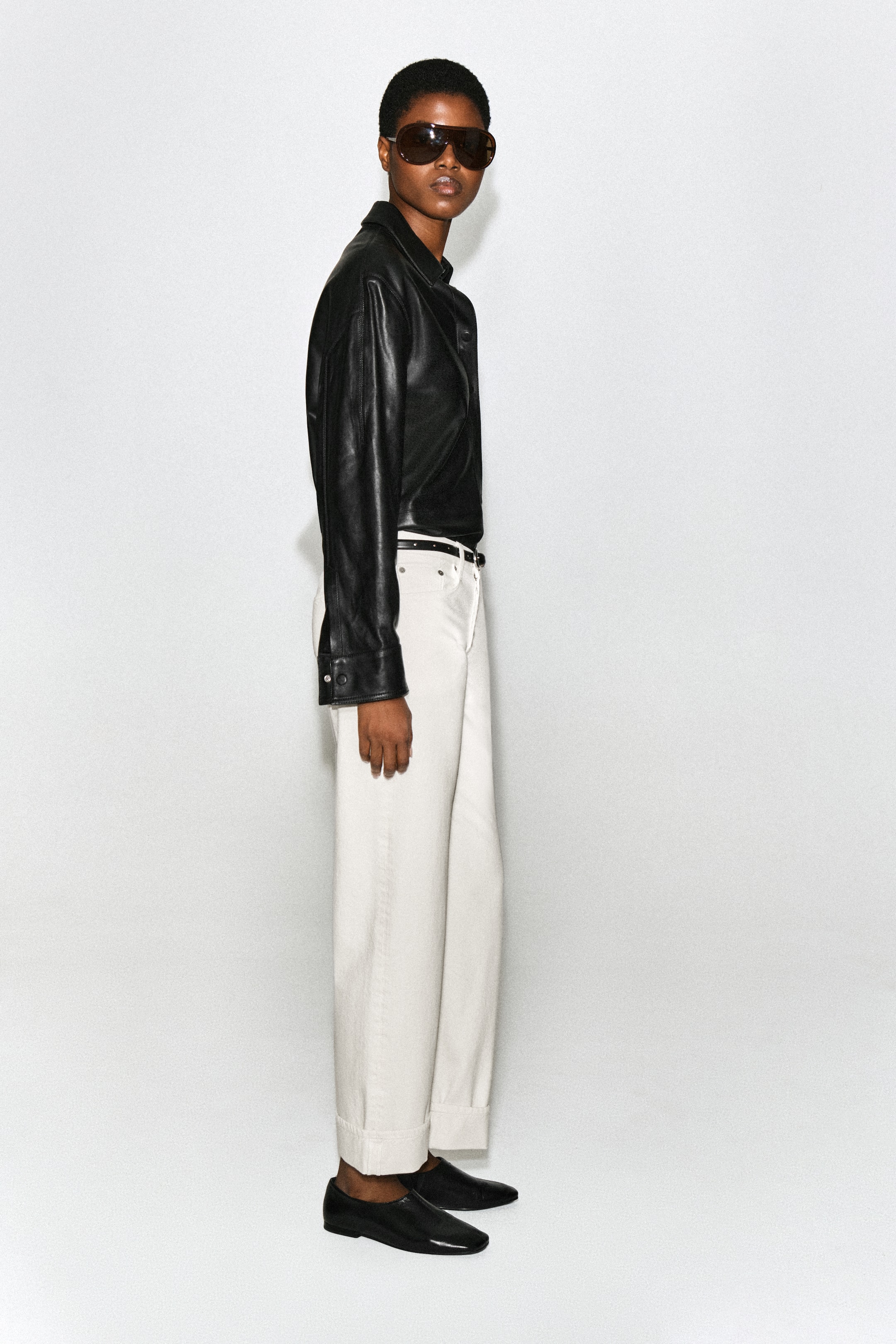 Grotere afbeelding bekijken: De FOLDE turn-up jeans met gebogen pijpen in offwhite worden gedragen door het model, dat in profiel staat. De jeans met hoge taille hebben een gebogen silhouet met brede omgeslagen broekspijpen en zijn gestyled met een dunne zwarte riem, een zwart leren overhemd en zwarte platte schoenen.