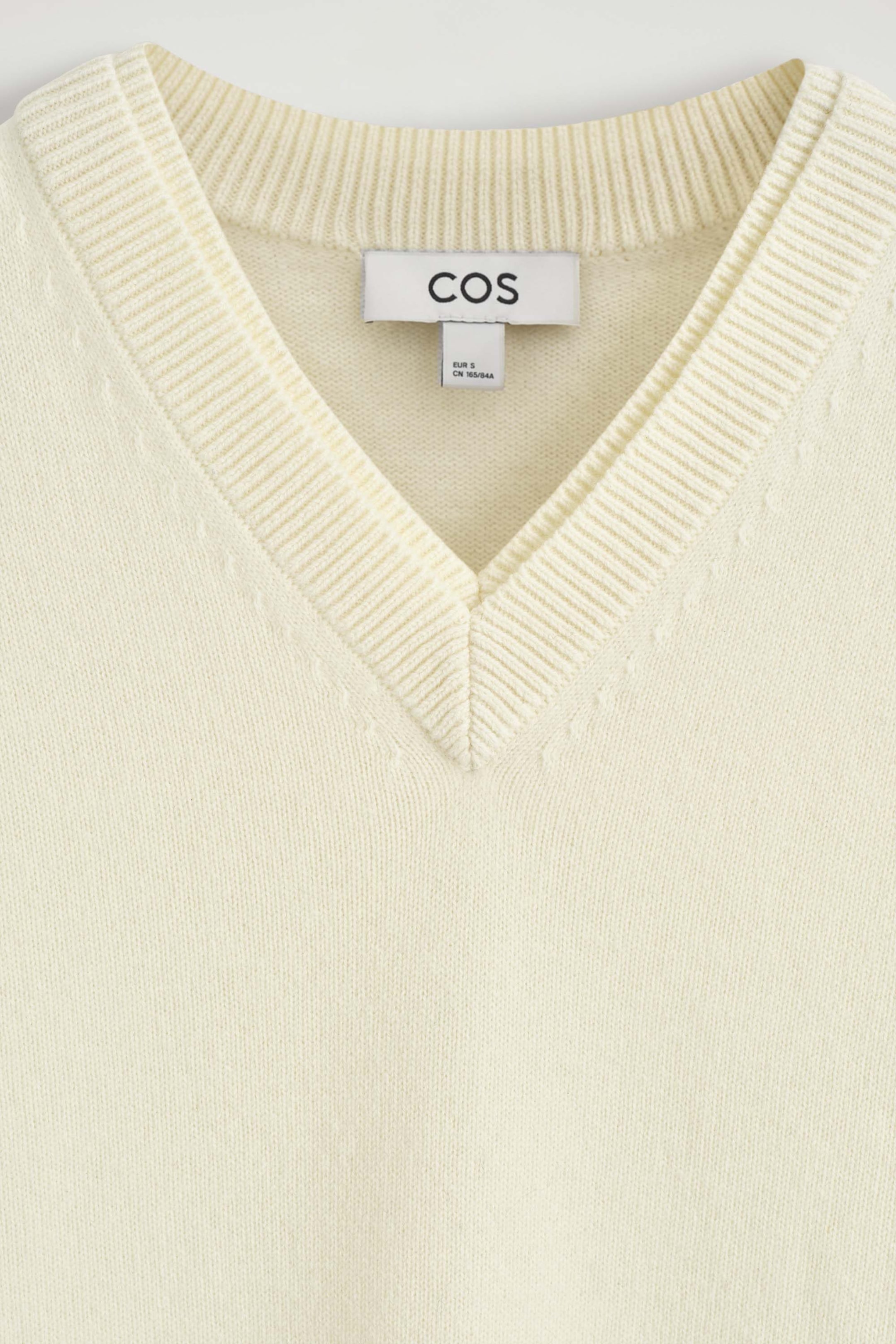 This close-up front view shows the cream-coloured Merino Wool Layered V-Neck Jumper, featuring a fine-knit texture and a double-layered collar design with a ribbed crew neck layered beneath a ribbed V-neck. This close-up front view shows the cream-coloured Merino Wool Layered V-Neck Jumper, featuring a fine-knit texture and a double-layered collar design with a ribbed crew neck layered beneath a ribbed V-neck.