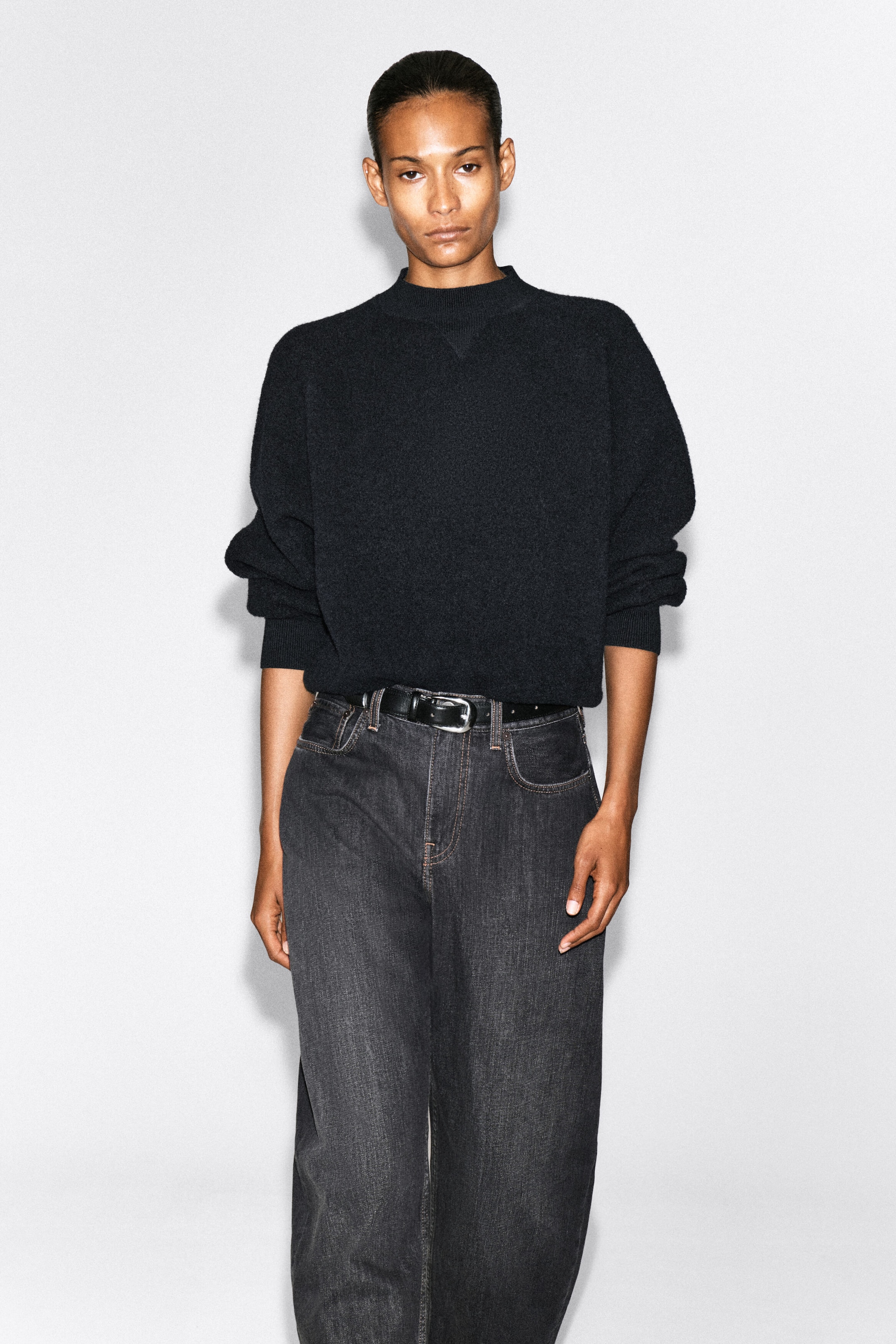 The model is wearing a black mock-neck knit sweater with a relaxed fit, long sleeves with ribbed cuffs, and a subtle triangular detail at the collar. The sweater is loosely tucked into dark gray, wide-leg denim jeans, styled with a black leather belt. The model is wearing a black mock-neck knit sweater with a relaxed fit, long sleeves with ribbed cuffs, and a subtle triangular detail at the collar. The sweater is loosely tucked into dark gray, wide-leg denim jeans, styled with a black leather belt.