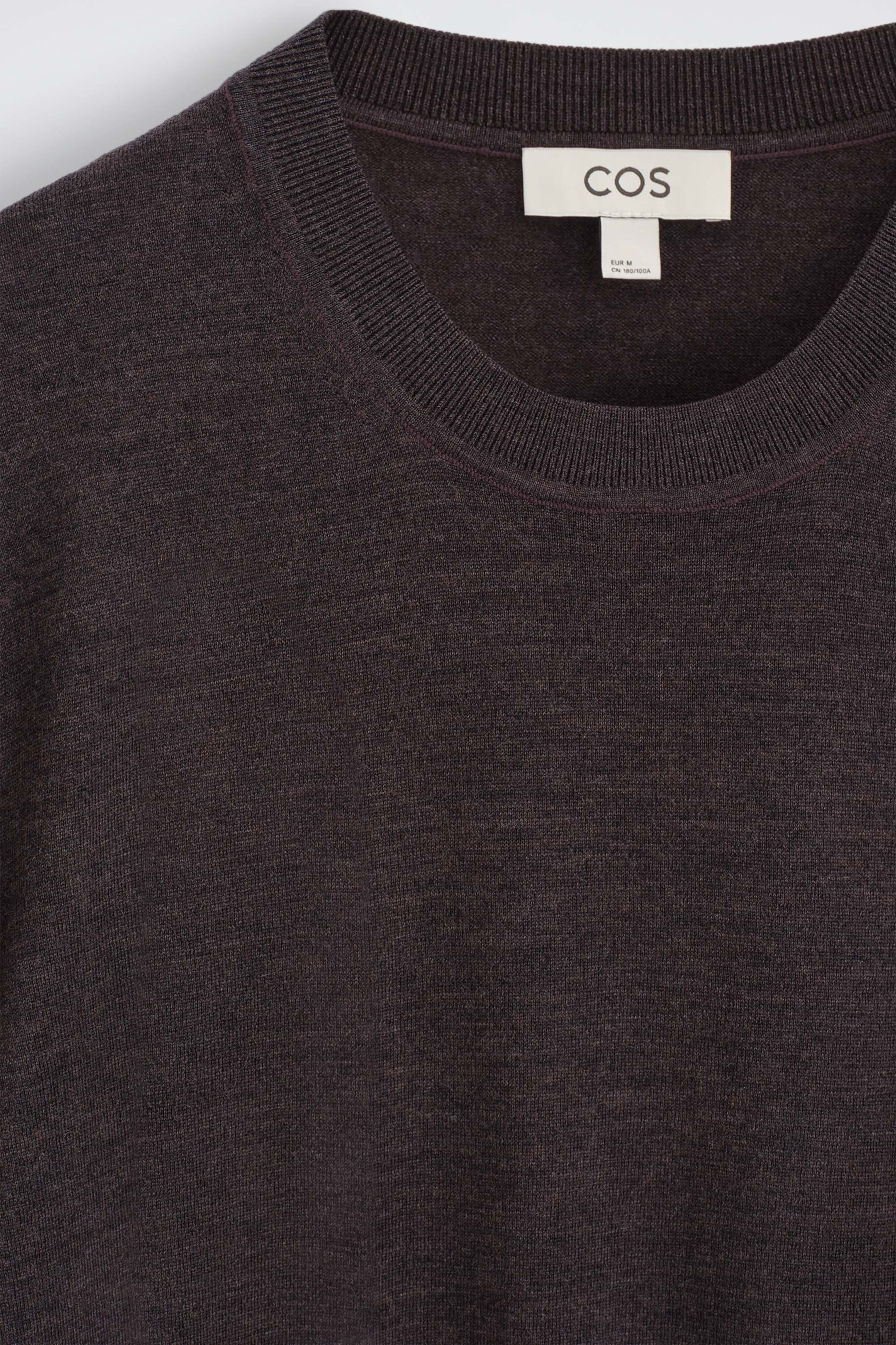 The slim merino wool crew-neck sweater is shown in a close-up front view, highlighting its dark brown melange fine-knit texture and ribbed crew neck. A white brand label is visible on the inside collar. The slim merino wool crew-neck sweater is shown in a close-up front view, highlighting its dark brown melange fine-knit texture and ribbed crew neck. A white brand label is visible on the inside collar.