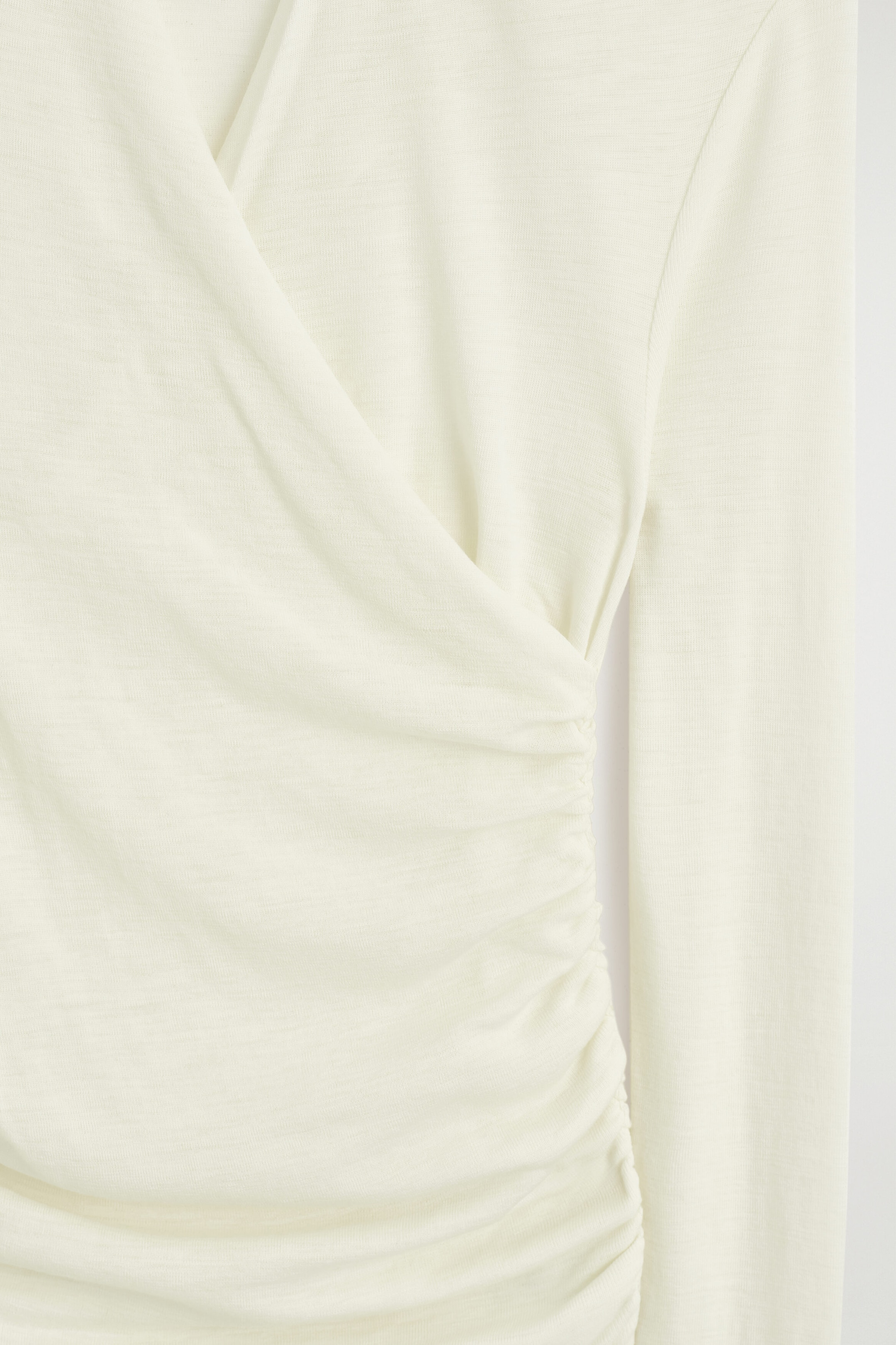 A cream-coloured wrap top is shown from the front, featuring a draped crossover design that gathers into ruched detailing along the side seam. The close-up highlights the fine-knit texture of the fabric and a long sleeve. A cream-coloured wrap top is shown from the front, featuring a draped crossover design that gathers into ruched detailing along the side seam. The close-up highlights the fine-knit texture of the fabric and a long sleeve.