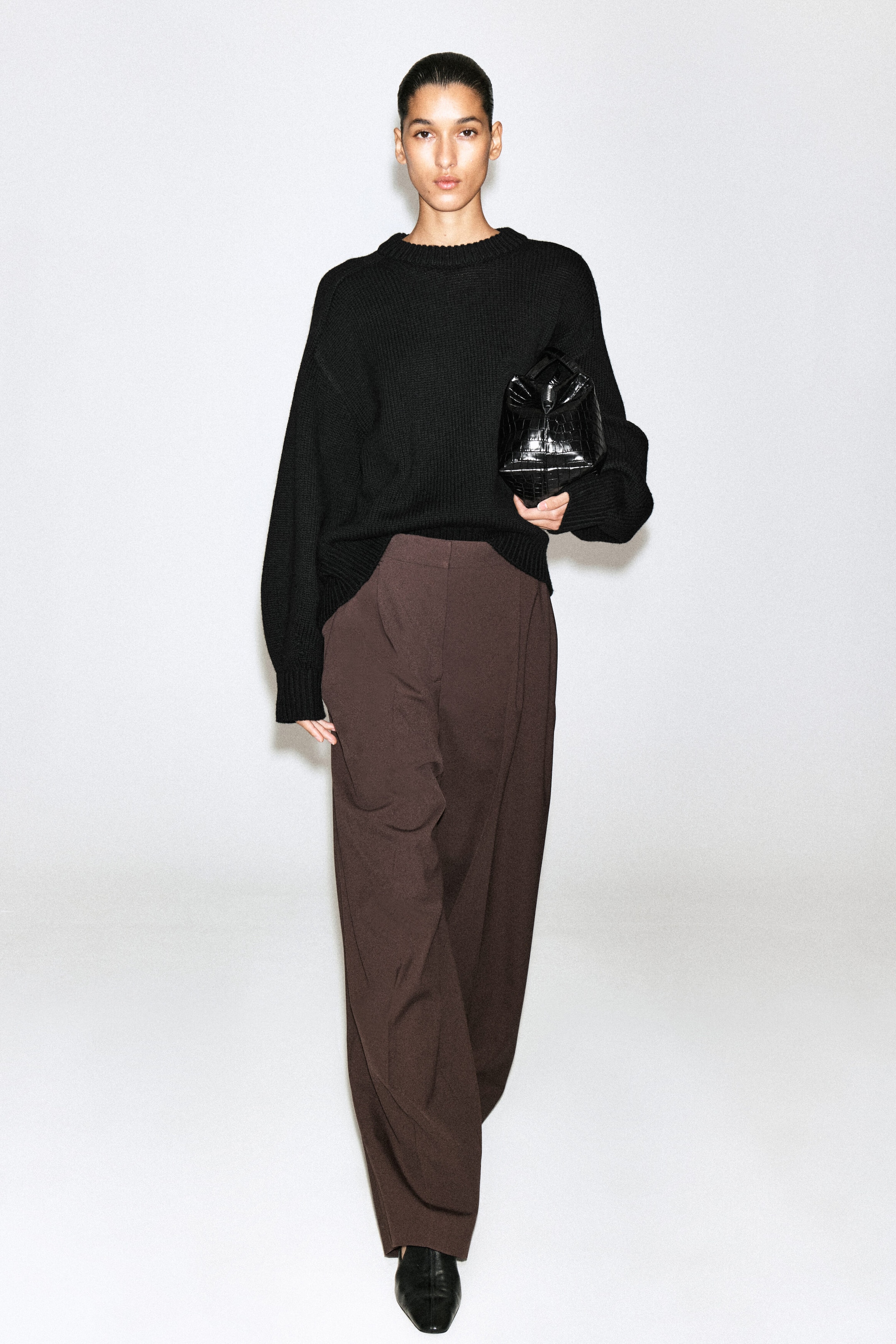 The black ribbed crew-neck jumper and brown wide-leg trousers are styled together in a front-facing view. The oversized jumper features dropped shoulders and long sleeves with ribbed cuffs, while the high-waisted, pleated trousers drape in a relaxed, wide-leg silhouette.