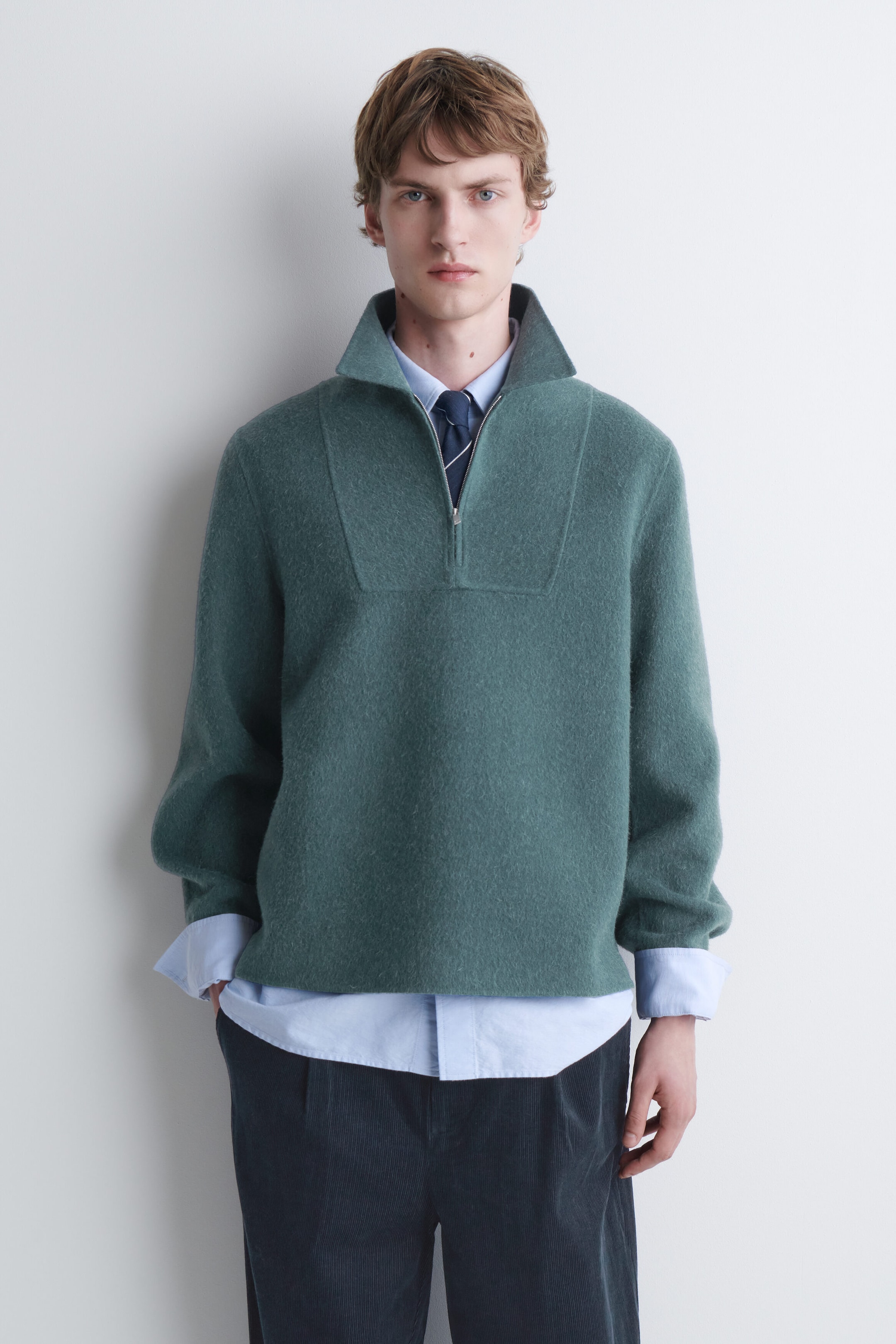 A green half-zip knit sweater is being worn by the model over a light blue collared shirt and a dark tie. The sweater features a high, open collar and is styled with dark trousers. A green half-zip knit sweater is being worn by the model over a light blue collared shirt and a dark tie. The sweater features a high, open collar and is styled with dark trousers.