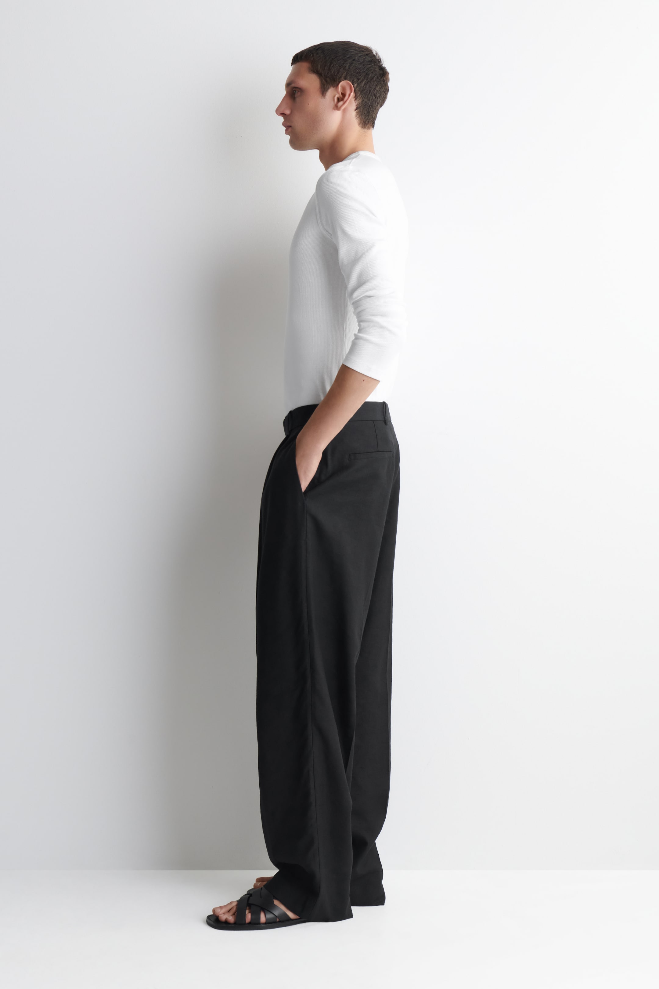 Größeres Bild ansehen: Schwarze Hose mit lockerer, weit geschnittener Silhouette, von der Seite gesehen. Eine Hand ruht in einer Seitentasche, und der Stoff fällt bis zum Boden und bedeckt teilweise Sandalen.