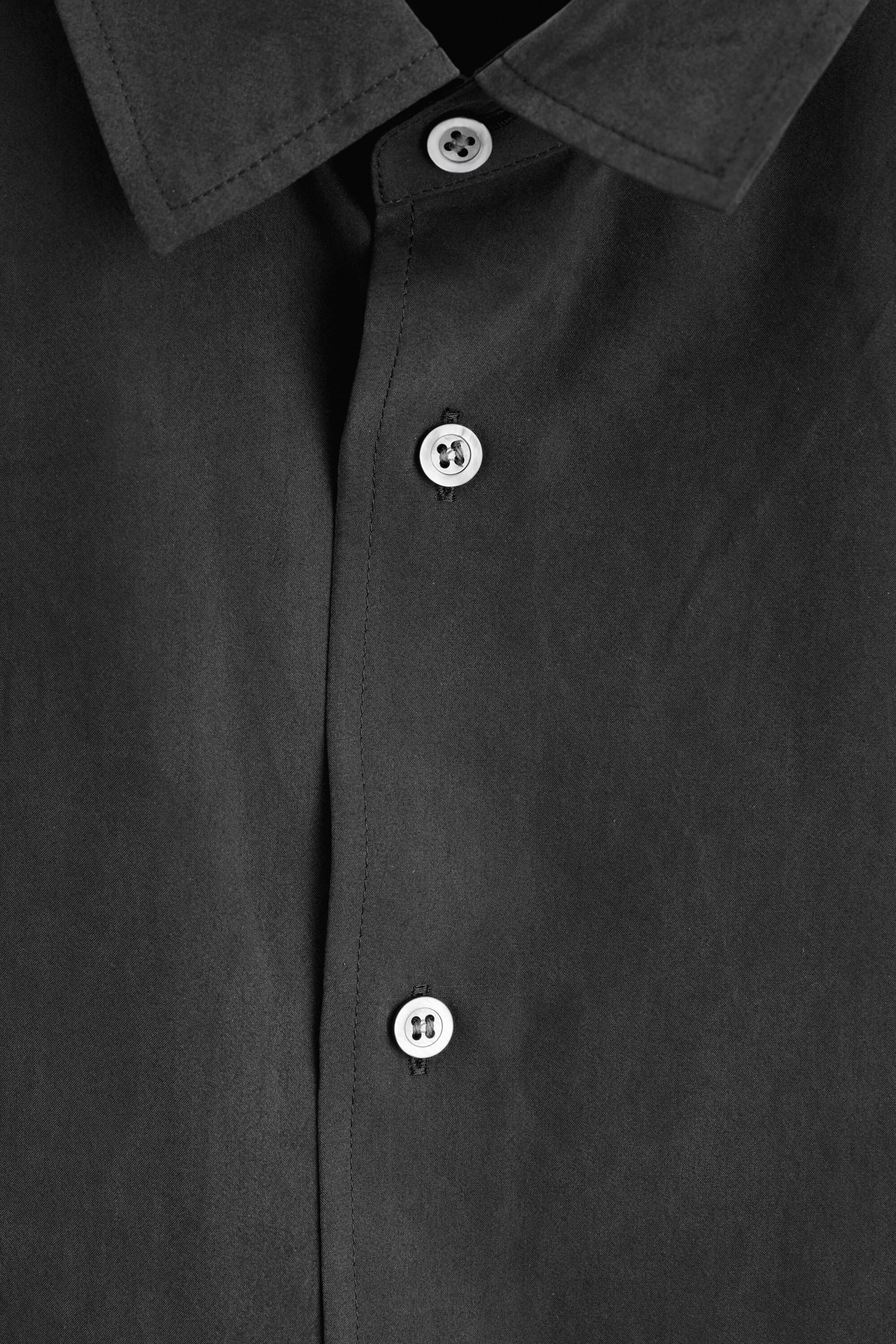 A close-up front view of a black sandwashed silk shirt showcases its matte, soft-textured fabric, a classic pointed collar, and a clean button-down placket. The shirt features contrasting light-coloured four-hole buttons with dark stitching along the front fastening. A close-up front view of a black sandwashed silk shirt showcases its matte, soft-textured fabric, a classic pointed collar, and a clean button-down placket. The shirt features contrasting light-coloured four-hole buttons with dark stitching along the front fastening.