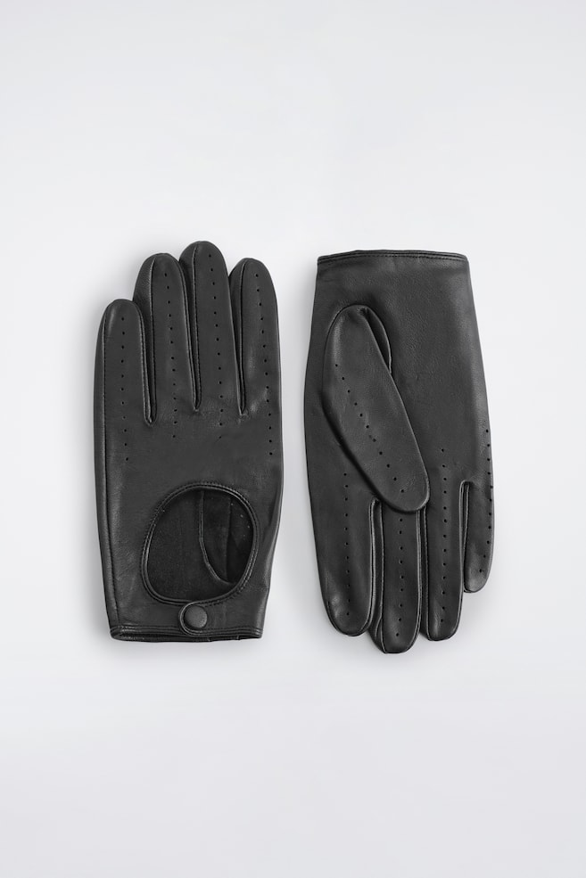 Une paire de gants de conduite en cuir noir est posée à plat, le gant gauche orienté de face pour mettre en valeur le design sur le dos de la main, avec une découpe circulaire, une fermeture à pression recouverte de cuir et des rangées de détails perforés le long des doigts. Le gant droit présente la paume, mettant en avant le pouce replié et des détails perforés assortis. - 1