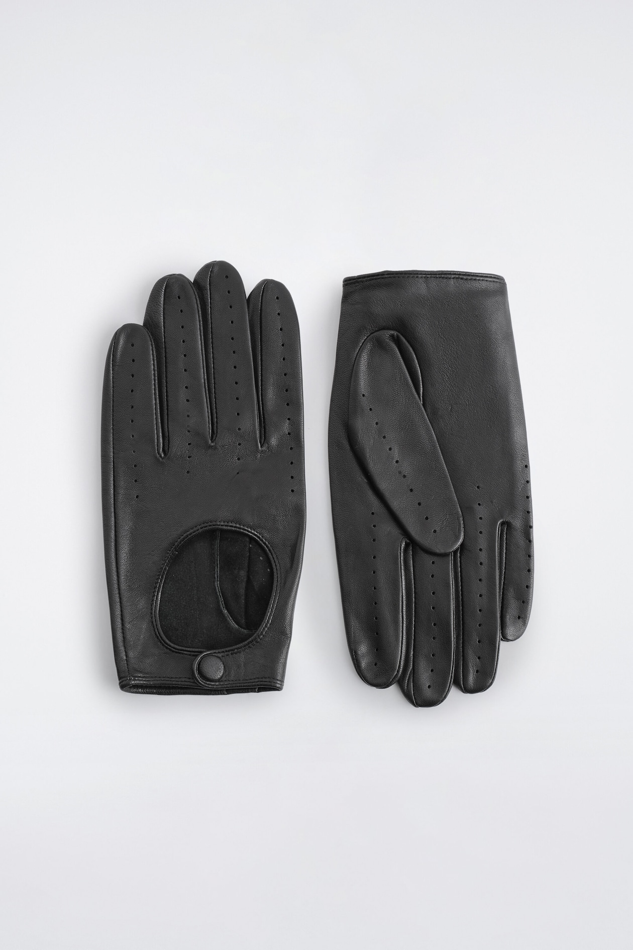 A pair of black leather driving gloves lies flat, with the left glove facing front to show the back-of-hand design featuring a circular cut-out, a leather-covered press-stud fastening, and rows of perforated detailing along the fingers. The right glove is shown palm side up, highlighting the folded thumb and matching perforated details.
