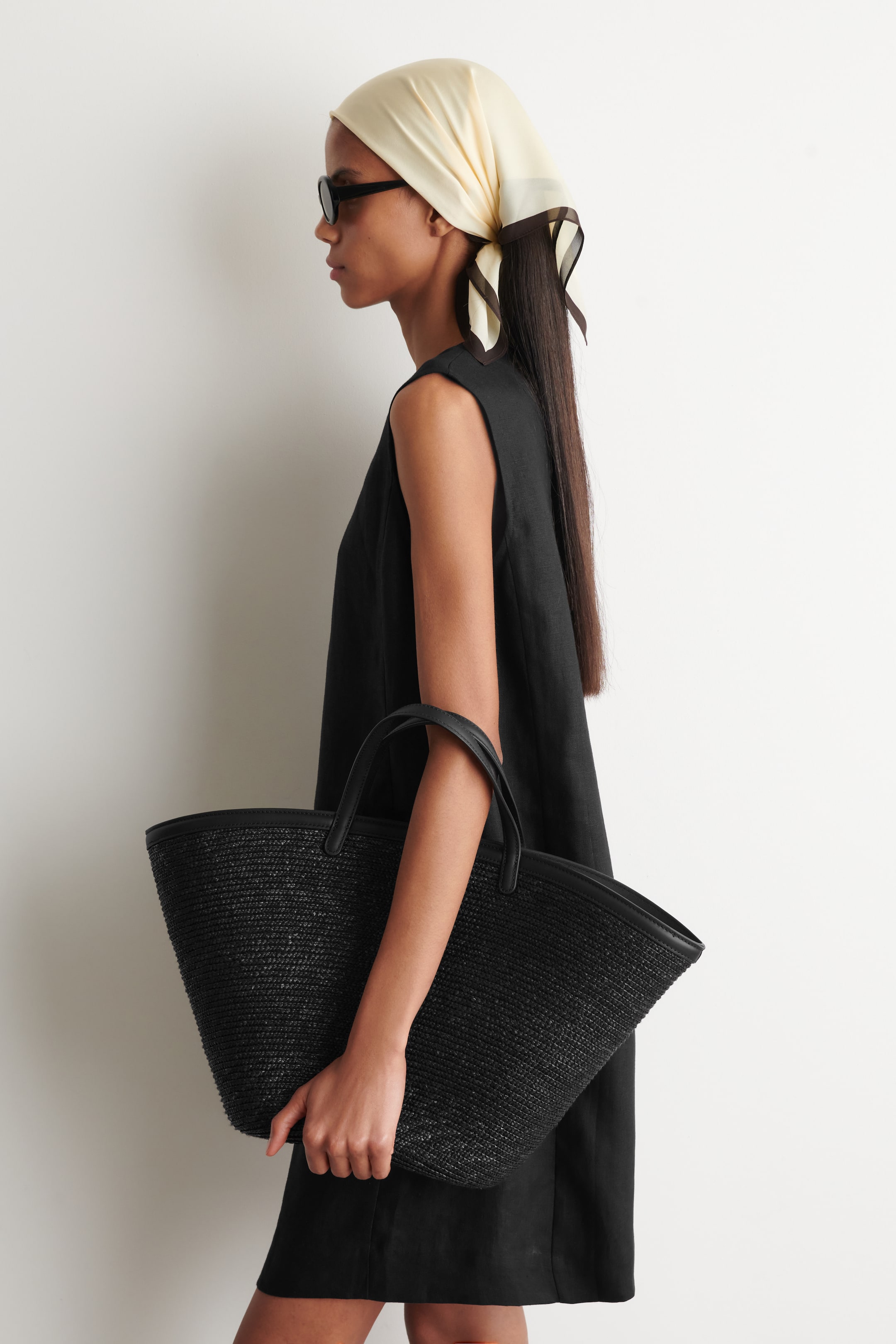 Agrandir l'image: Un sac fourre-tout noir tissé est tenu par le côté, affichant sa silhouette effilée, sa texture semblable au raphia noir, ses poignées supérieures lisses et sa bordure assortie le long du bord supérieur.