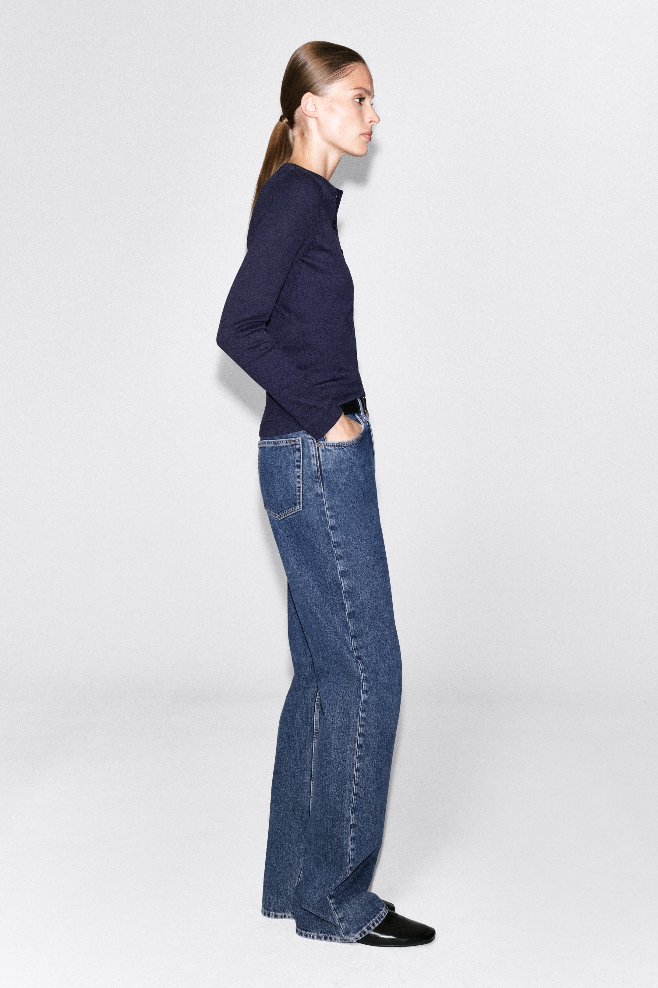 The model is wearing a navy blue ribbed cotton henley top, featuring a slim fit, long sleeves, and a buttoned neckline placket. The top is paired with blue jeans, with the model standing in profile and resting one hand in a front pocket. The model is wearing a navy blue ribbed cotton henley top, featuring a slim fit, long sleeves, and a buttoned neckline placket. The top is paired with blue jeans, with the model standing in profile and resting one hand in a front pocket.