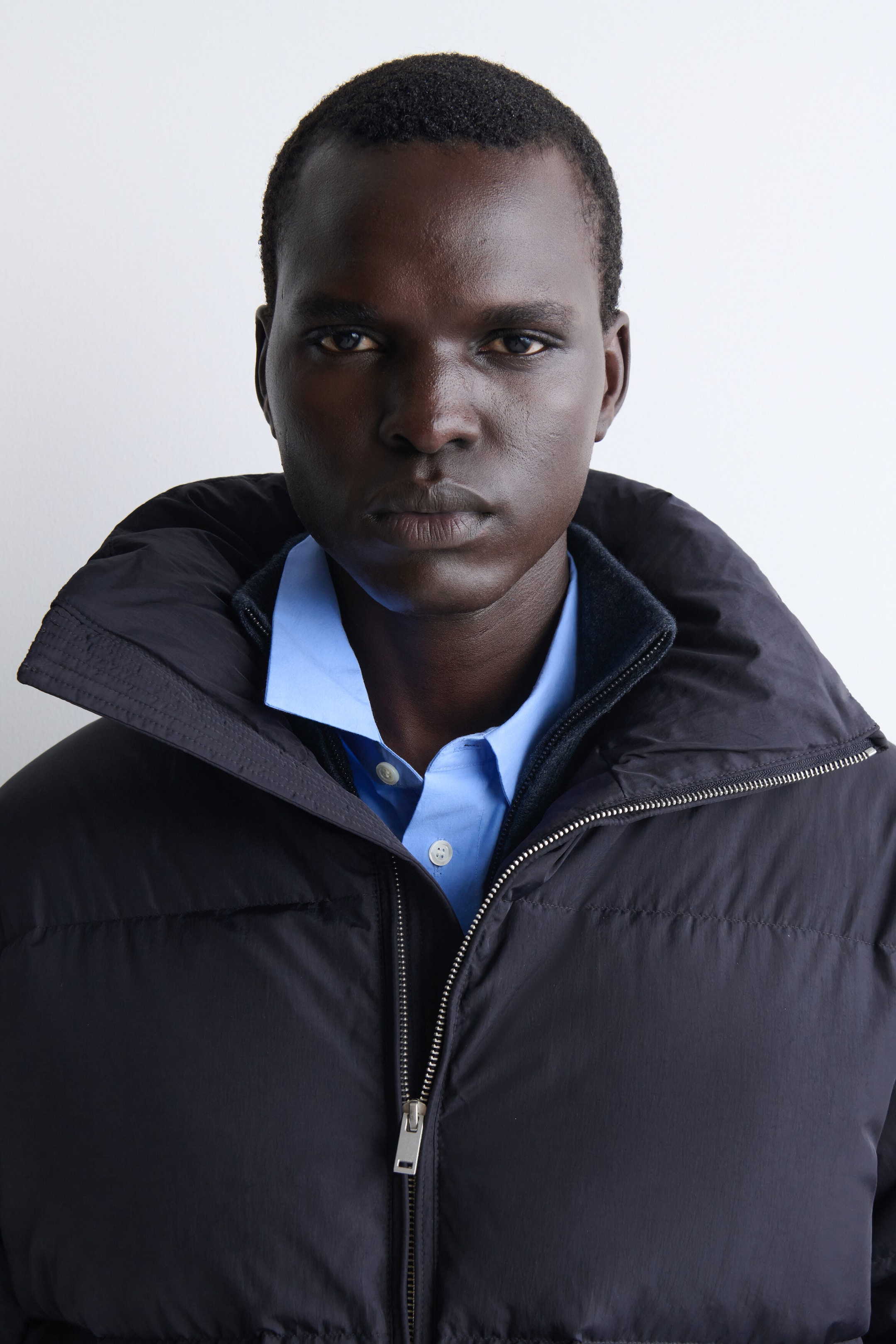 This dark navy funnel-neck nylon down puffer jacket is shown from a front-facing, close-up angle, featuring a high, padded collar flared open with a silver front zipper. The jacket is layered over a dark mock-neck knit and a light blue button-down shirt.