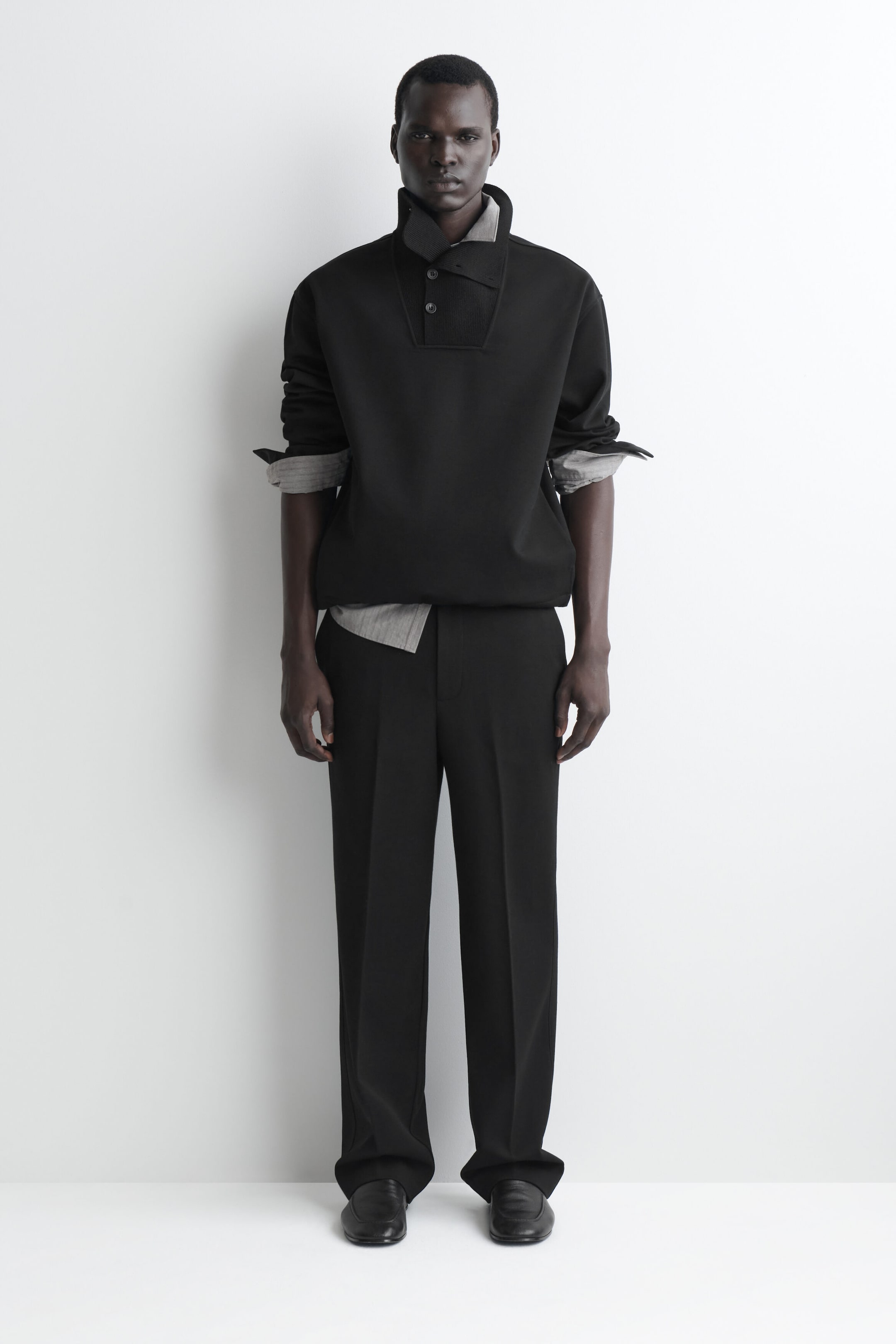 A black high-neck pullover and matching straight-leg trousers are shown from the front, styled with a gray collared shirt layered underneath. The pullover features a high collar with an asymmetrical button placket and pushed-up sleeves, while the trousers drape in a relaxed fit with a central crease. A black high-neck pullover and matching straight-leg trousers are shown from the front, styled with a gray collared shirt layered underneath. The pullover features a high collar with an asymmetrical button placket and pushed-up sleeves, while the trousers drape in a relaxed fit with a central crease.