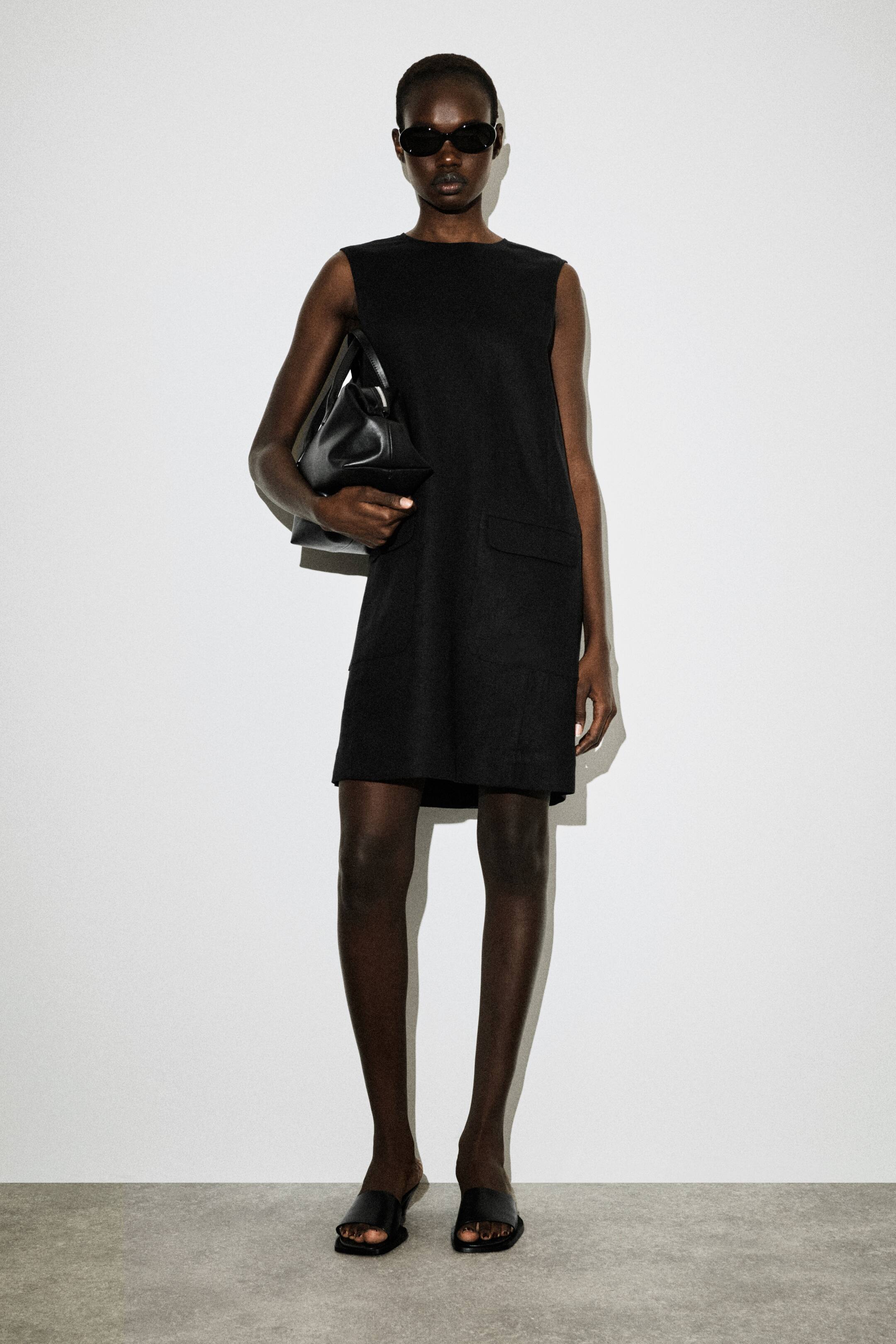 הצגת תמונה גדולה יותר: שמלת מיני שחורה מוצגת מהחזית, עם מפתח עגול ועיצוב ללא שרוולים. שני כיסי טלאי מלבניים נראים בחזית החצאית, והשמלה בעלת גזרה רפויה וישרה שנופלת מעל הברכיים.