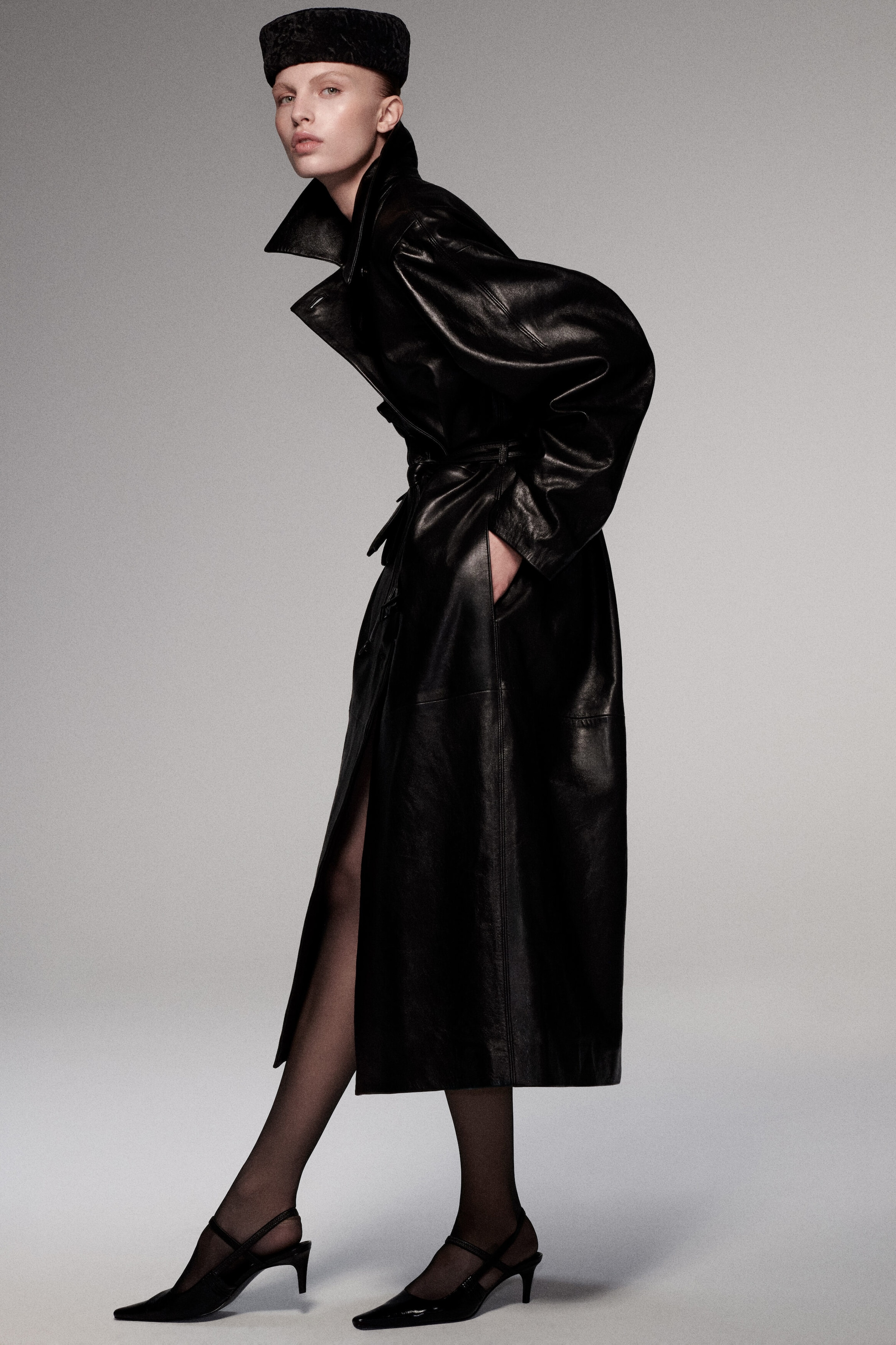 An oversized belted black leather long coat is presented in a three-quarter side profile, highlighting its voluminous, structured silhouette. The coat features a high pointed collar, oversized relaxed sleeves, a waist cinched with a matching leather tie belt, and a long calf-length hem. An oversized belted black leather long coat is presented in a three-quarter side profile, highlighting its voluminous, structured silhouette. The coat features a high pointed collar, oversized relaxed sleeves, a waist cinched with a matching leather tie belt, and a long calf-length hem.