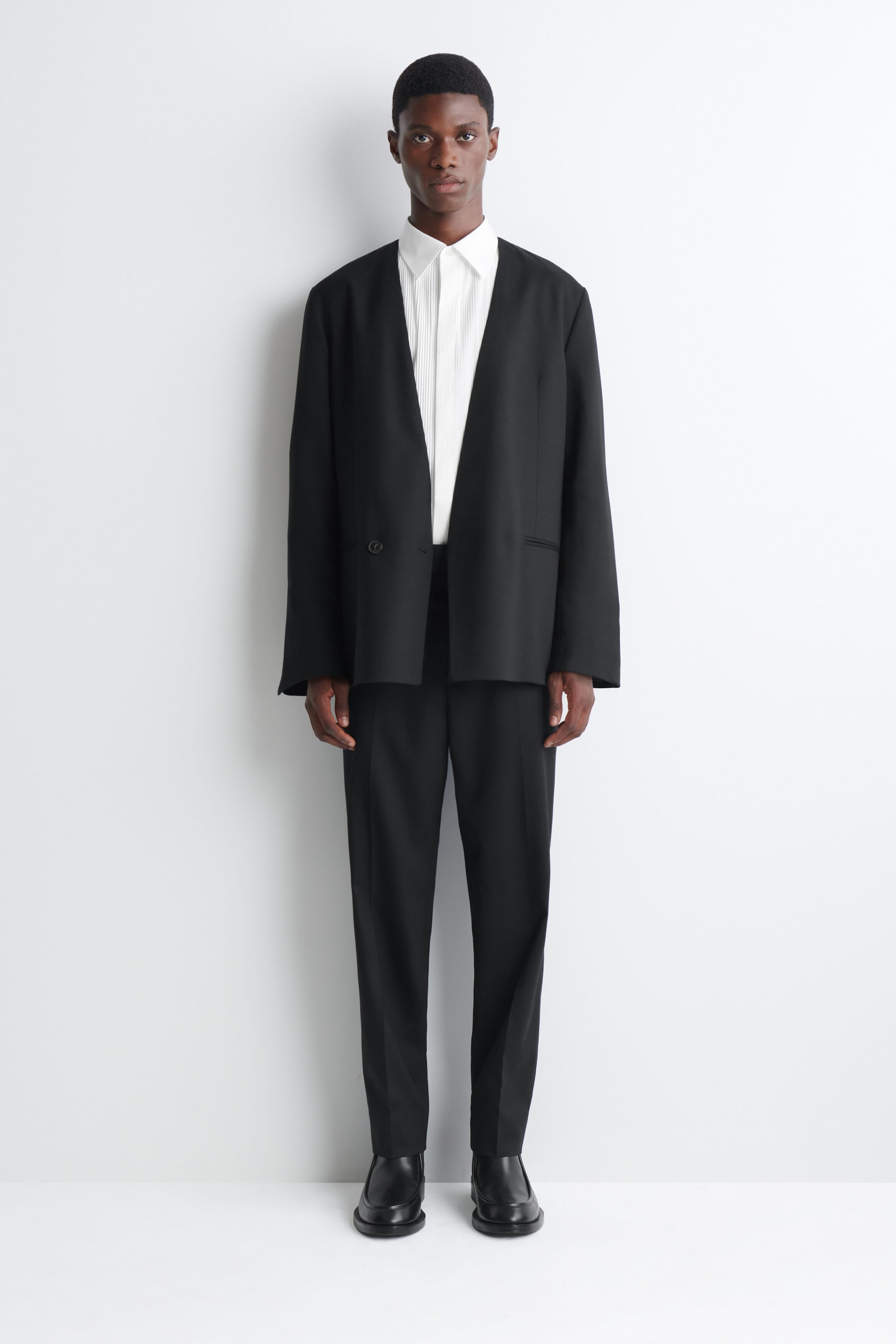 A black collarless suit, featuring a relaxed-fit blazer with a deep V-neck and matching straight-leg trousers, is worn by the model. The outfit is layered over a white pleated button-up shirt and styled with black leather shoes. A black collarless suit, featuring a relaxed-fit blazer with a deep V-neck and matching straight-leg trousers, is worn by the model. The outfit is layered over a white pleated button-up shirt and styled with black leather shoes.