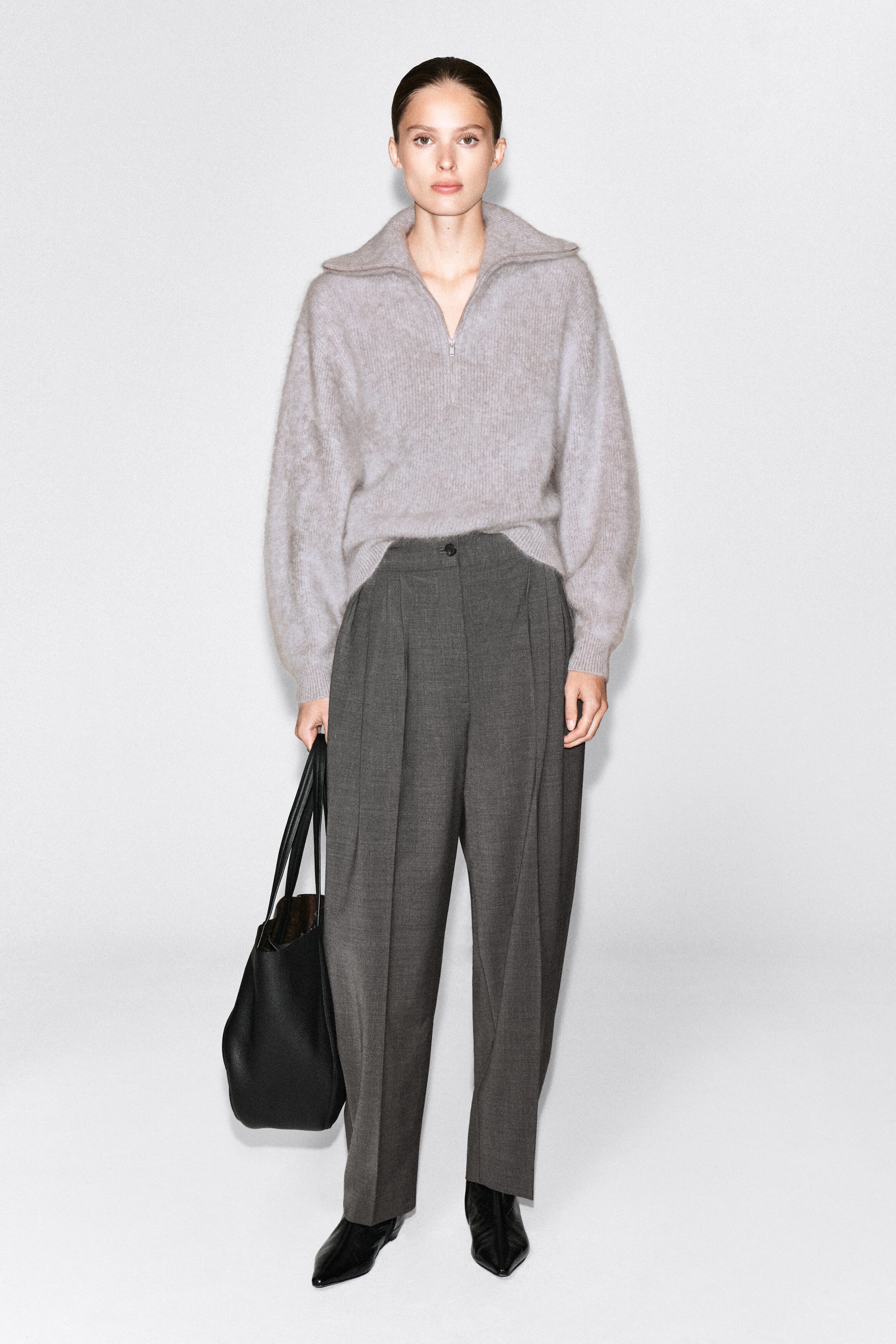 The model is wearing a light gray brushed cashmere sweater with a half-zip collar, tucked at the front into dark gray pleated trousers. The model is also holding a black leather tote bag. The model is wearing a light gray brushed cashmere sweater with a half-zip collar, tucked at the front into dark gray pleated trousers. The model is also holding a black leather tote bag.