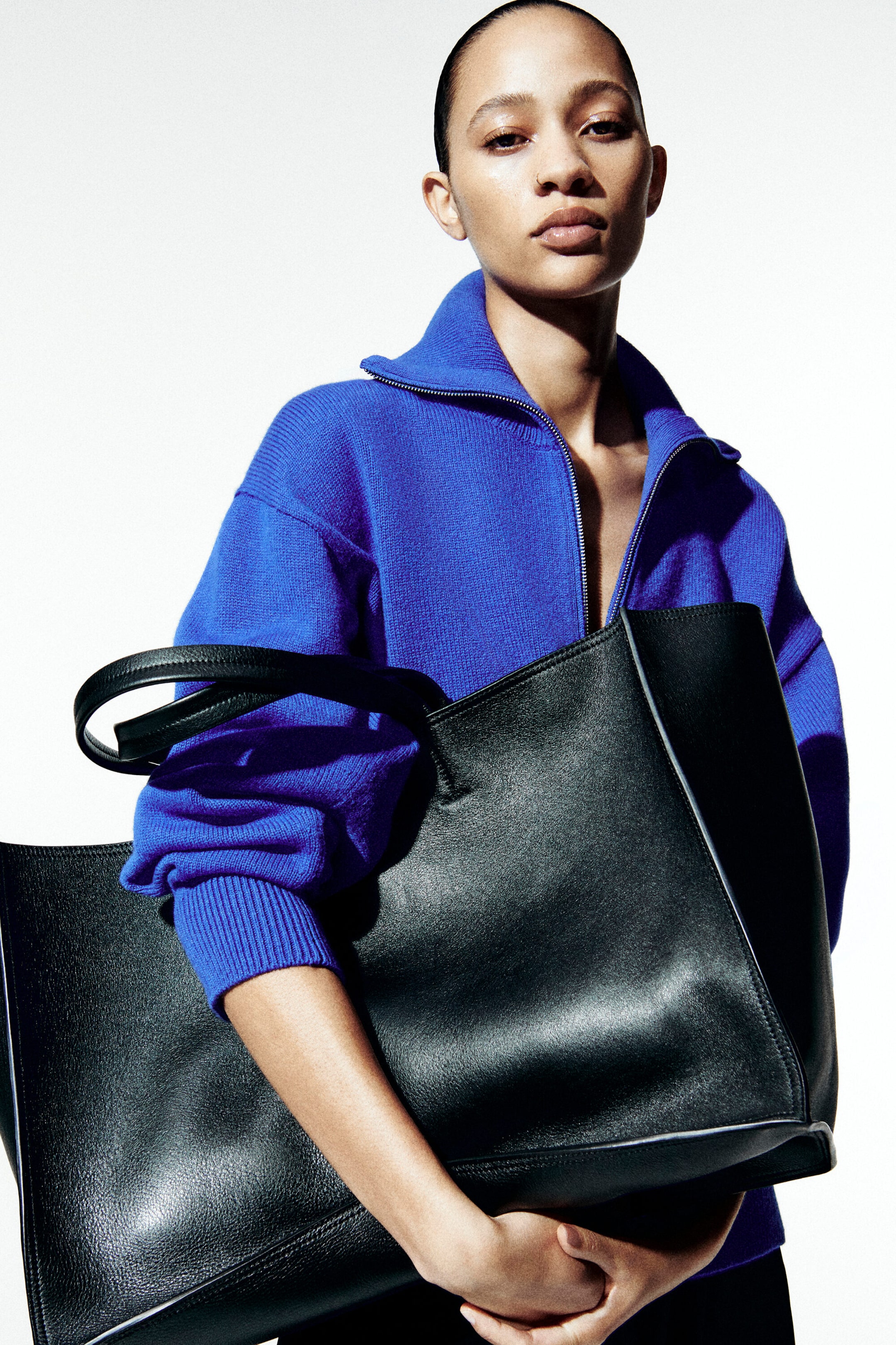 A model is wearing a bright blue, relaxed-fit knit jumper featuring a half-zip funnel neckline and ribbed cuffs while holding a large, structured black leather tote bag with dual shoulder straps across the front. A model is wearing a bright blue, relaxed-fit knit jumper featuring a half-zip funnel neckline and ribbed cuffs while holding a large, structured black leather tote bag with dual shoulder straps across the front.