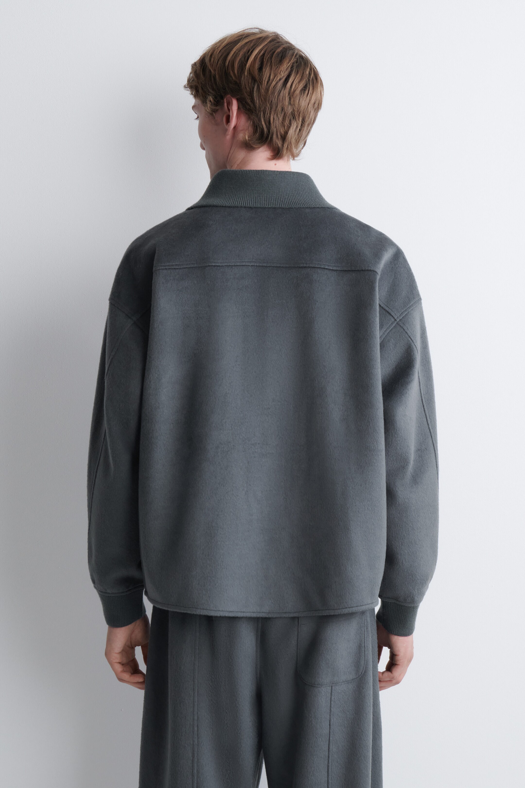 Une veste gris foncé à texture polaire, dotée d’un col montant côtelé, d’épaules tombantes et d’une empiècement au dos, est portée par le mannequin, vu de dos. La veste est assortie à un pantalon gris foncé. Une veste gris foncé à texture polaire, dotée d’un col montant côtelé, d’épaules tombantes et d’une empiècement au dos, est portée par le mannequin, vu de dos. La veste est assortie à un pantalon gris foncé.